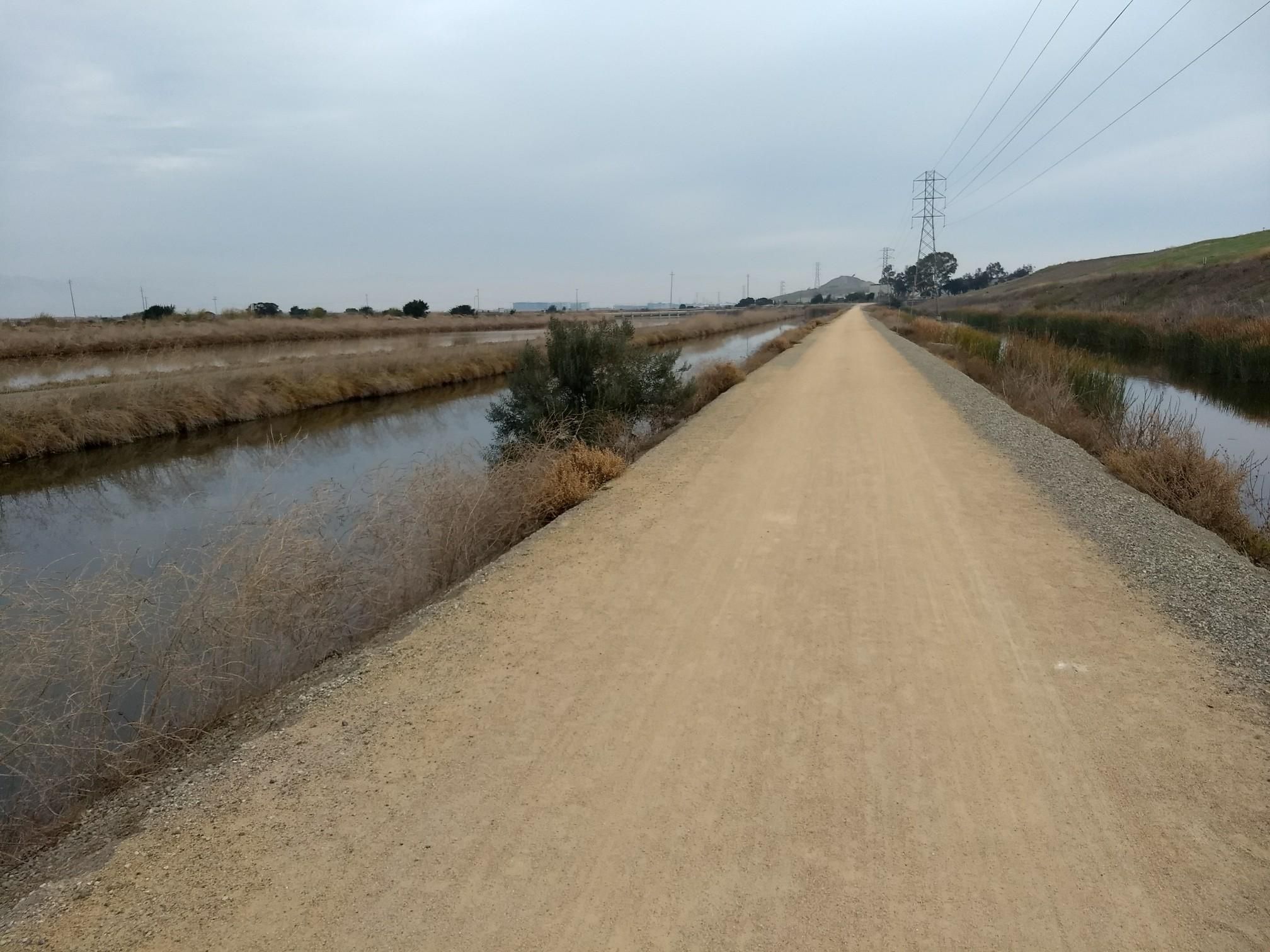 A dirt path runs between two water canals under an overcast sky. - San Francisco Bay Trail