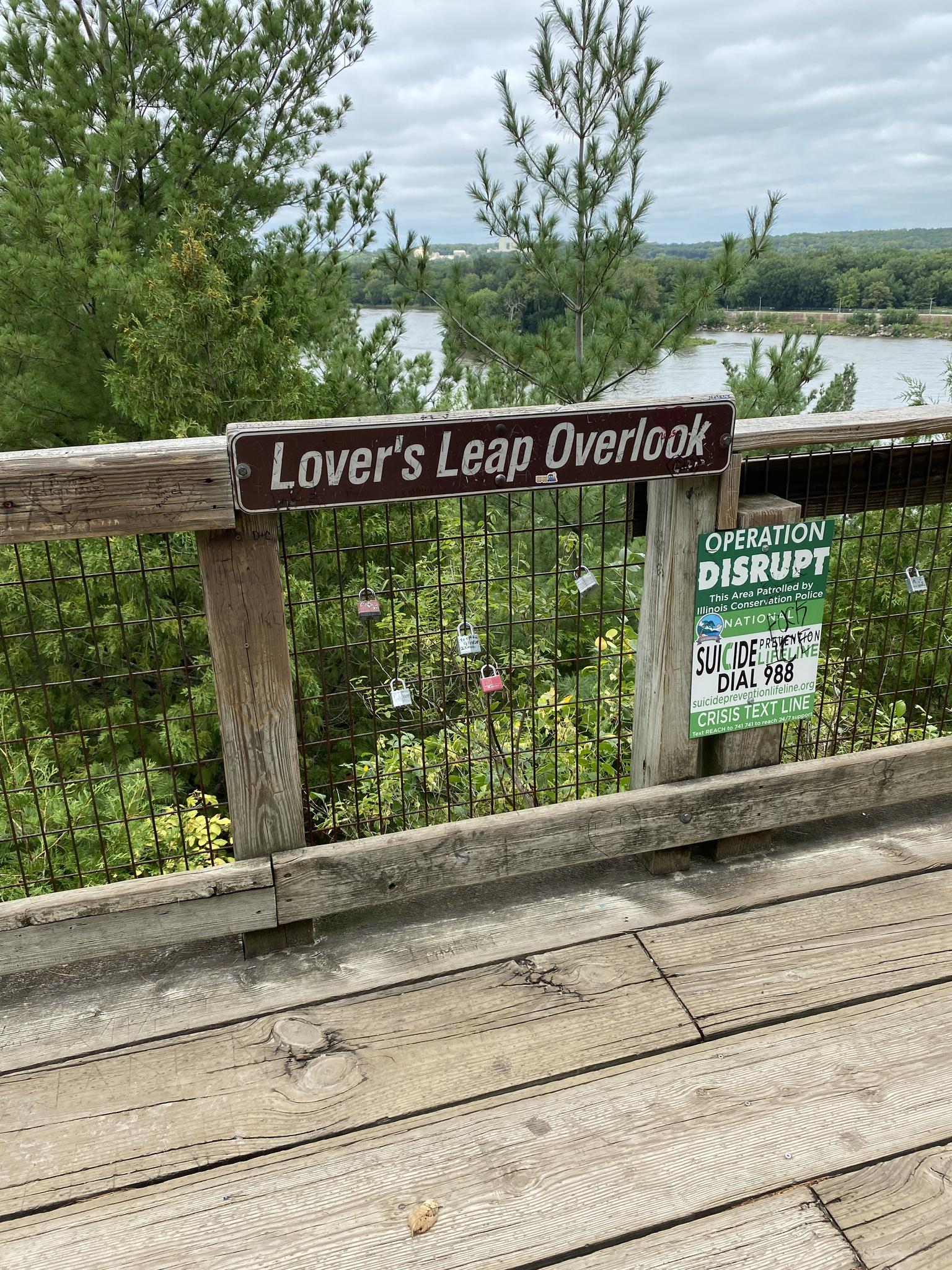 Overlook railing with signs and locks, calm river, green trees, overcast sky.