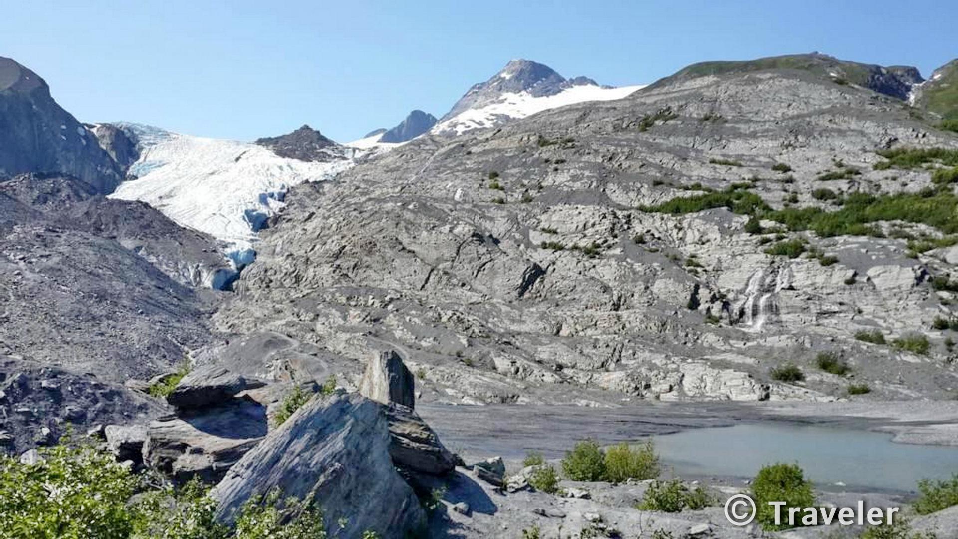 Bright view of a large glacier descending rocky mountains towards a small lake.