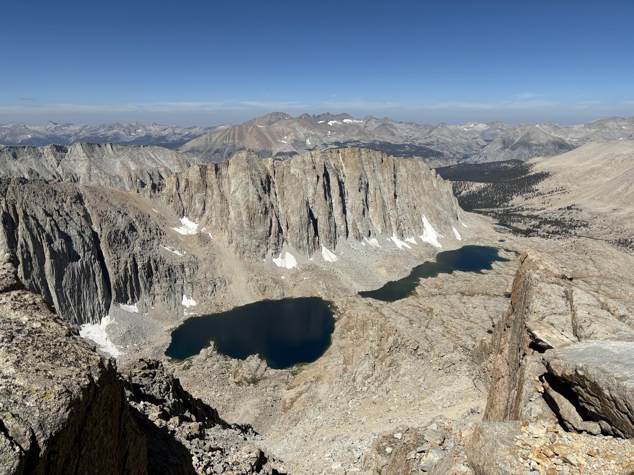 High-angle view of rugged mountains, two dark alpine lakes, and snow patches.