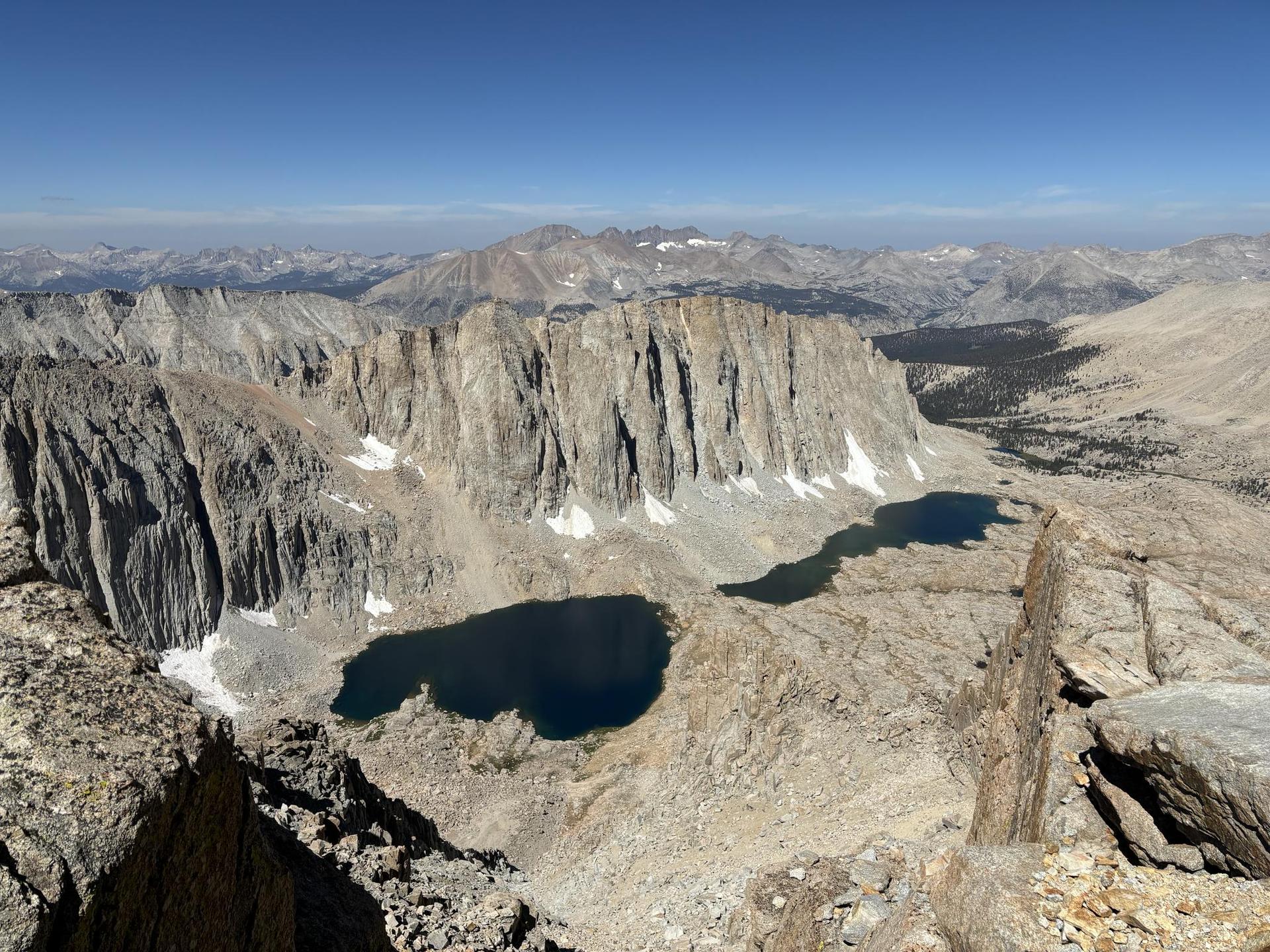 High-angle view of rugged mountains, two dark alpine lakes, and snow patches.