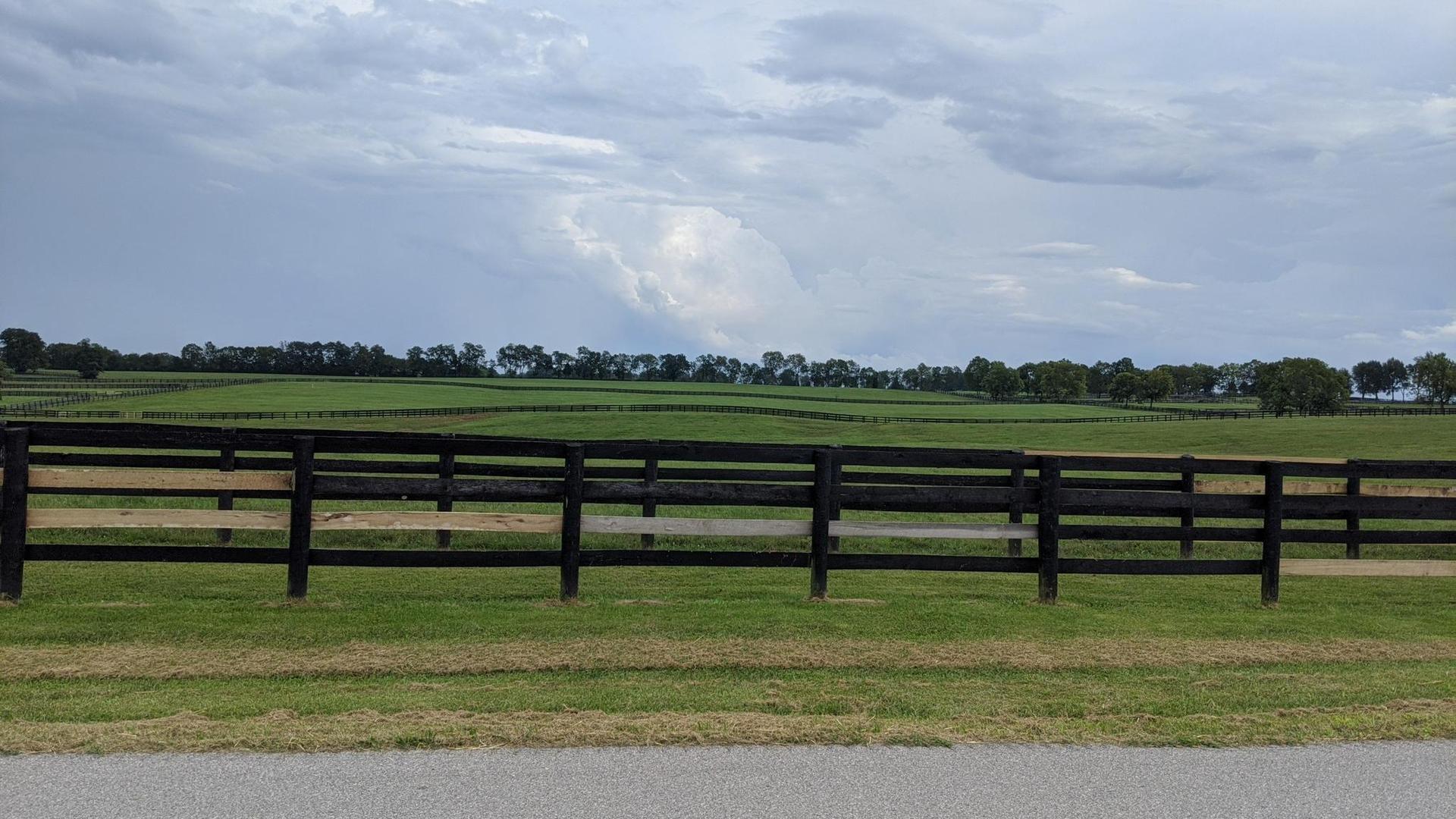 Black wooden fence in front of green fields under a dramatic cloudy sky.