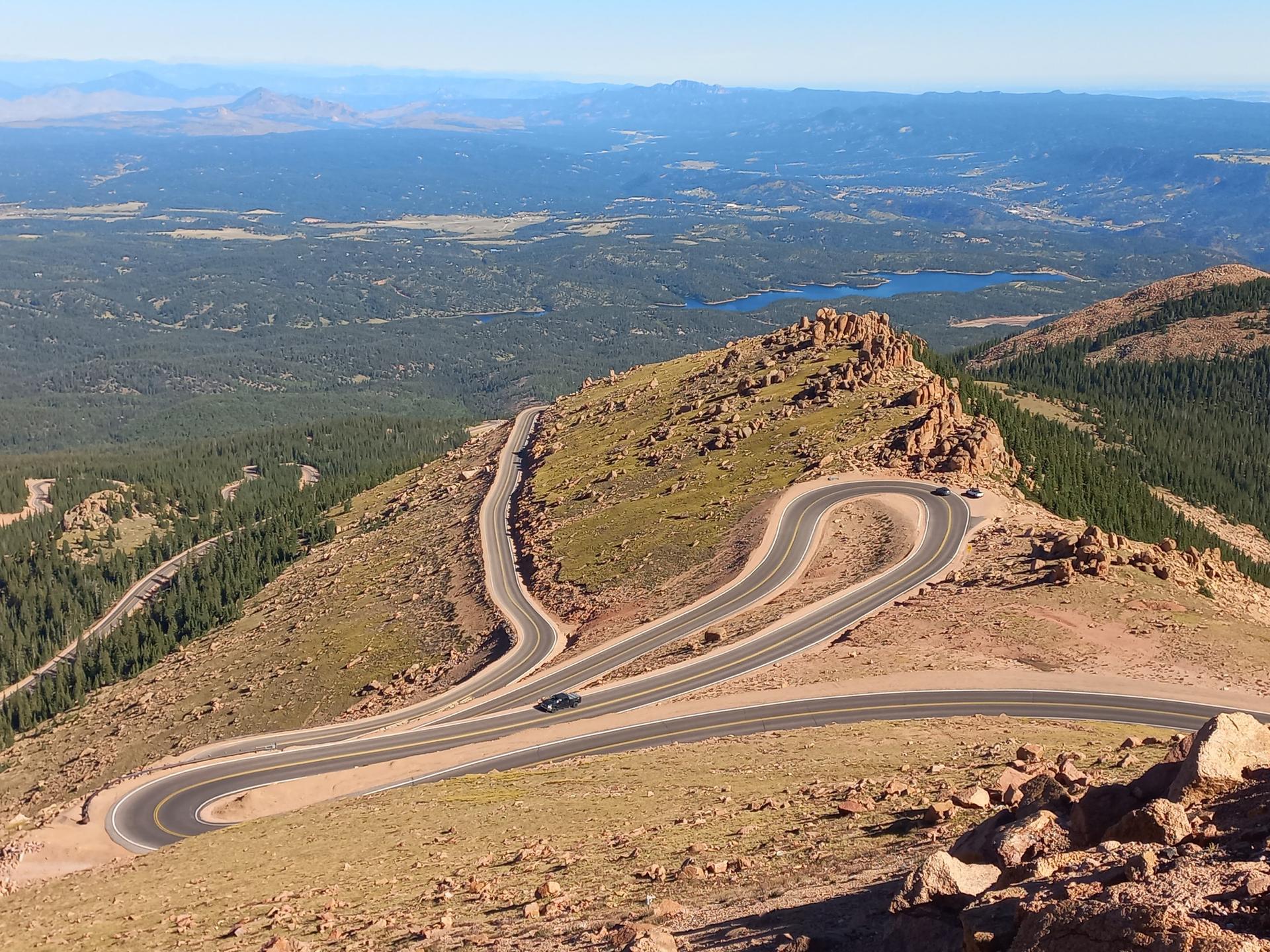 Winding switchback road on a mountain, with distant lakes and forests.