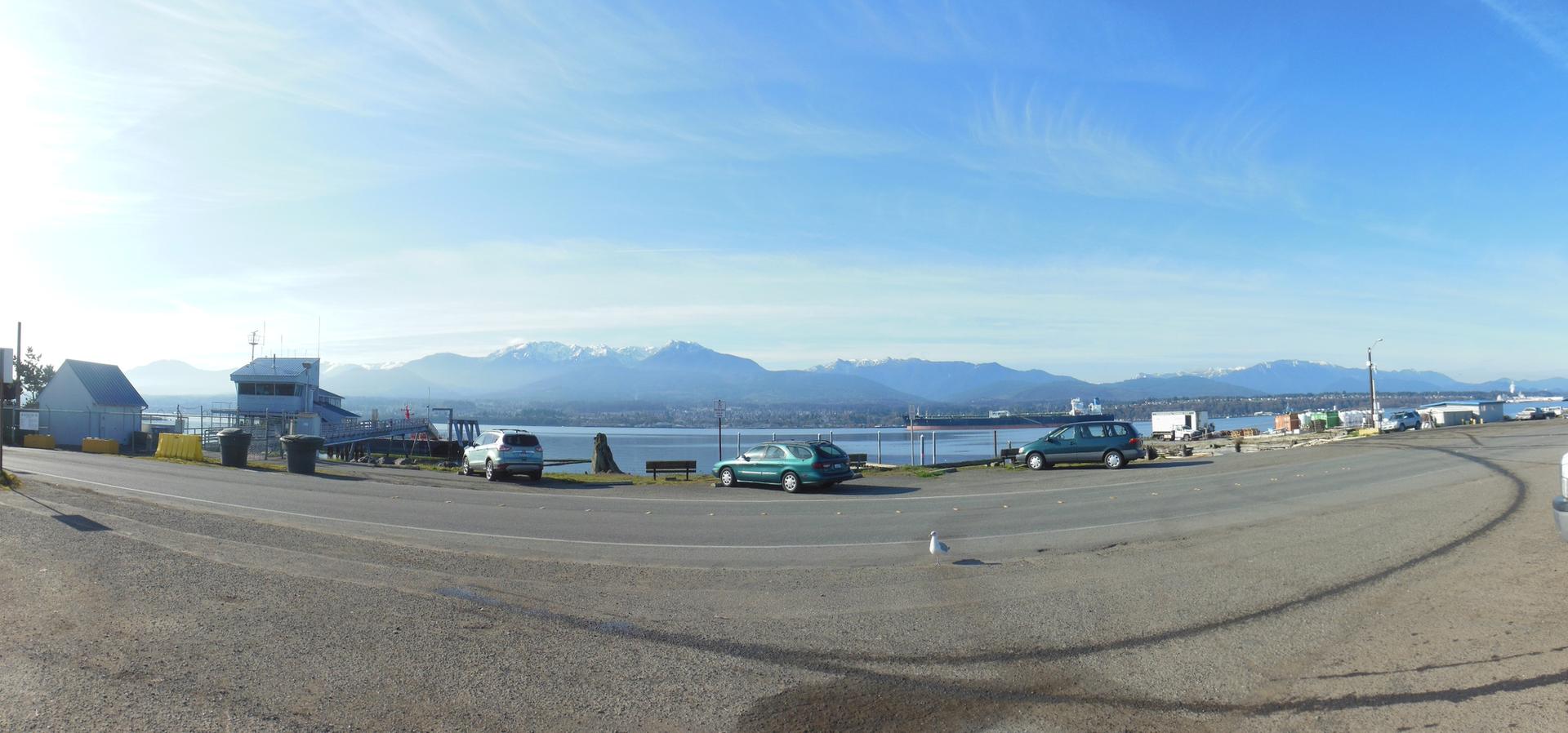 Panoramic view of a waterfront with parked cars, calm water, and distant mountains.