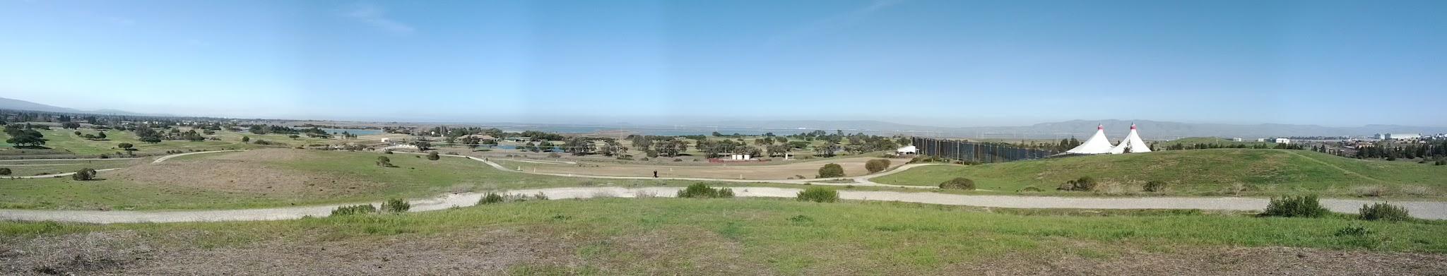Panoramic view of rolling green hills with distant structures under a clear blue sky.