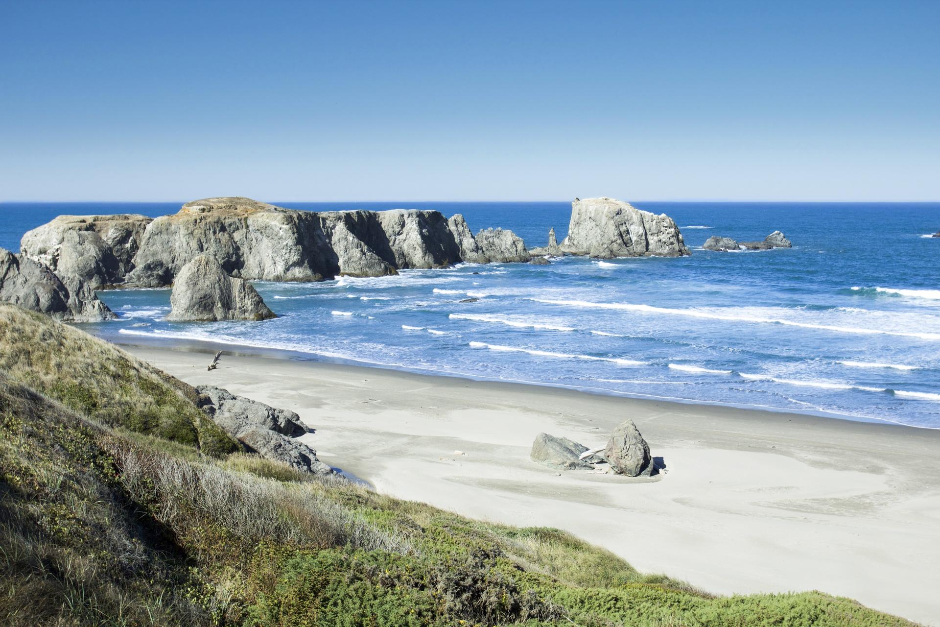 Sunny beach with large rock formations in the ocean under a clear sky.
