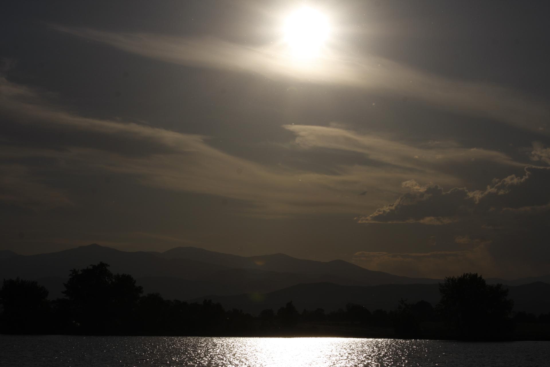 Sun glares over water, silhouetting distant mountains and trees at dusk.