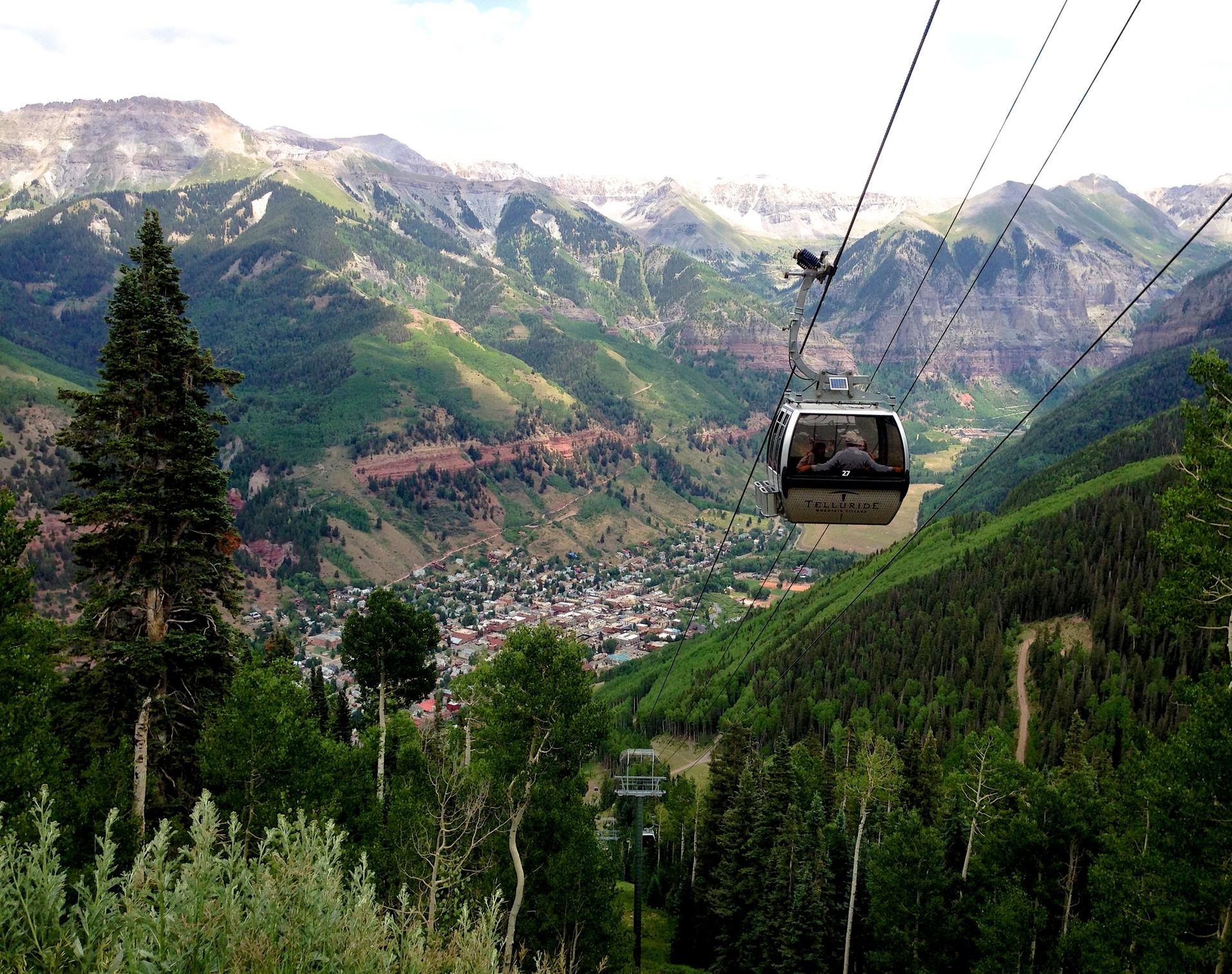 Gondola car with passengers ascends over a green mountain valley and town.