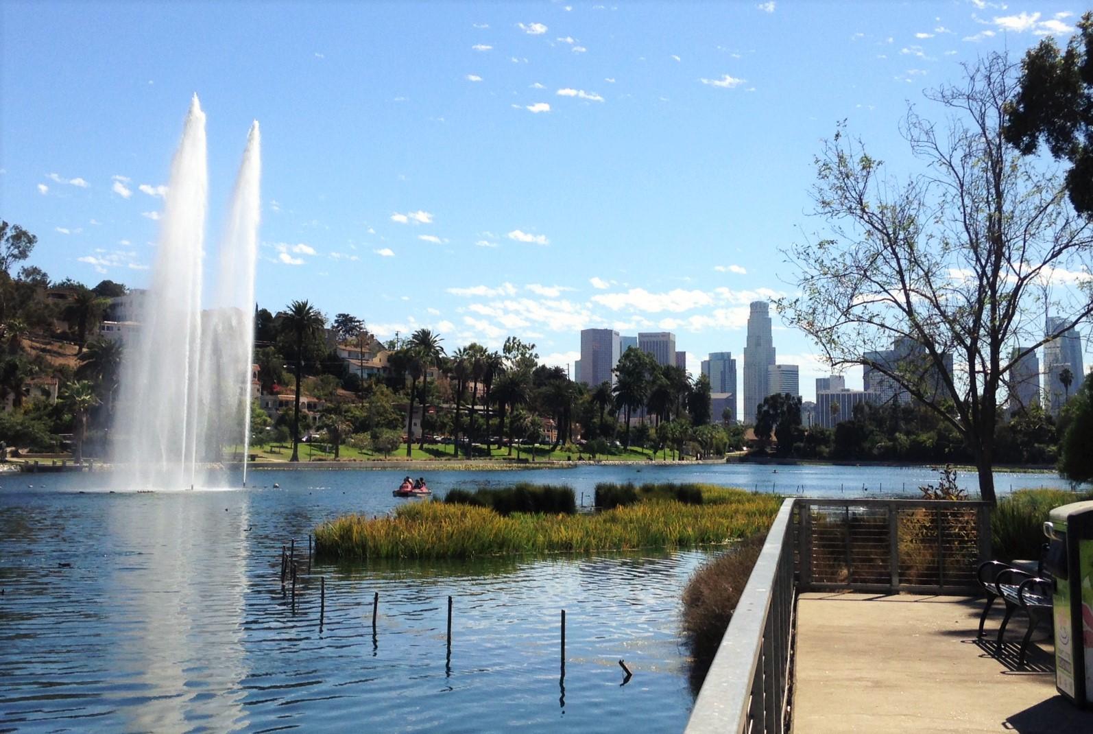 Sunny lake scene with fountains, distant city, and people boating.
