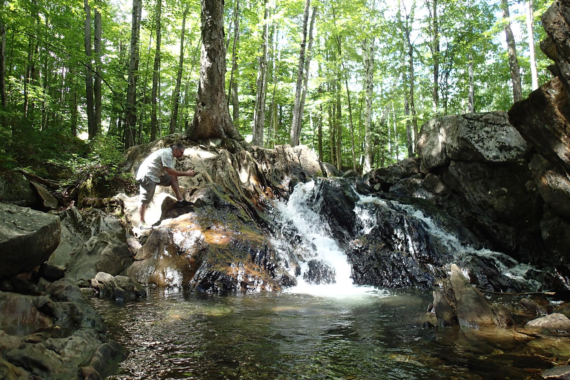 Person crouching by a small waterfall in a sunlit, rocky forest.