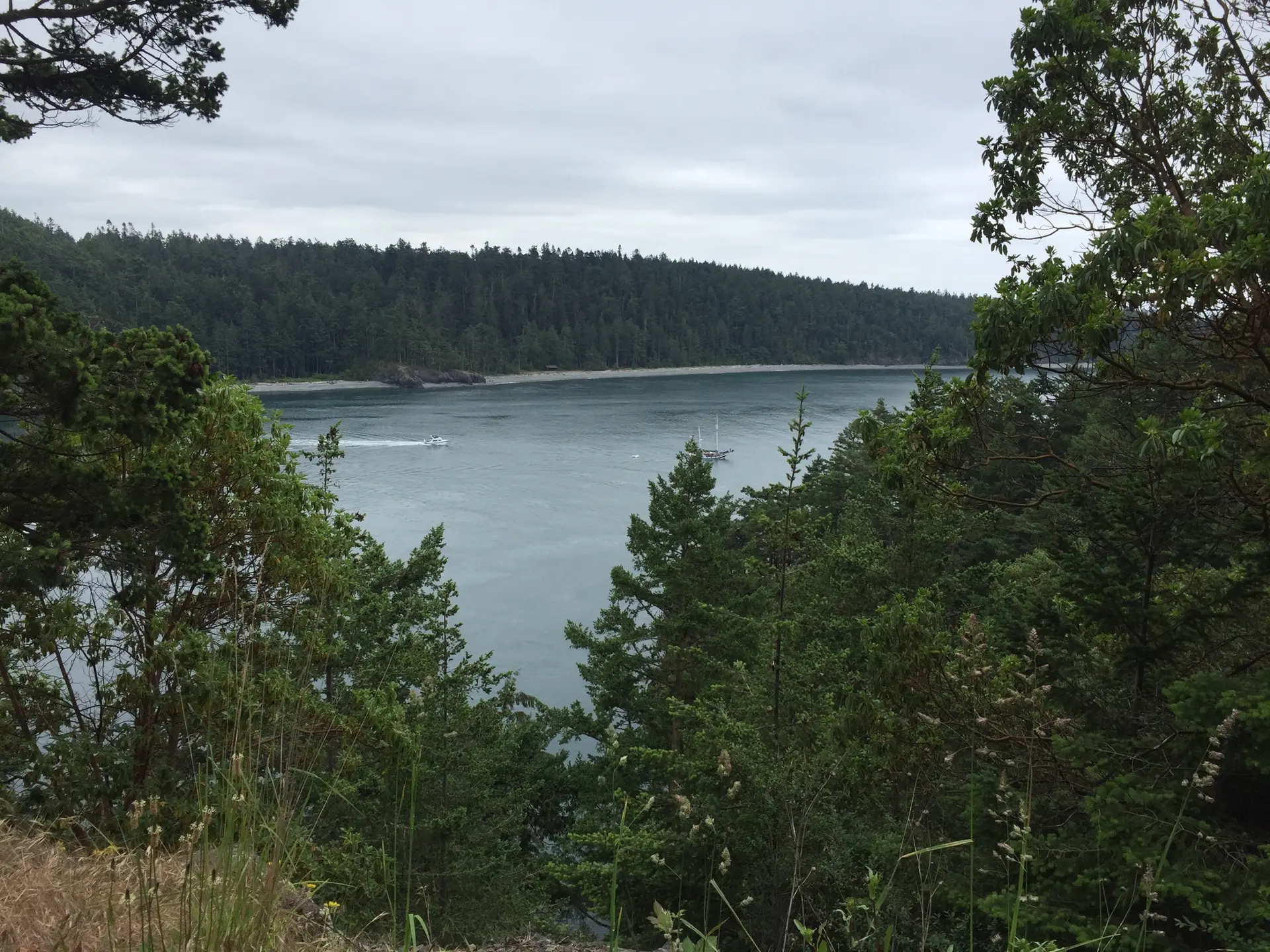 Overcast view of a wide bay with distant forested land and small boats.
