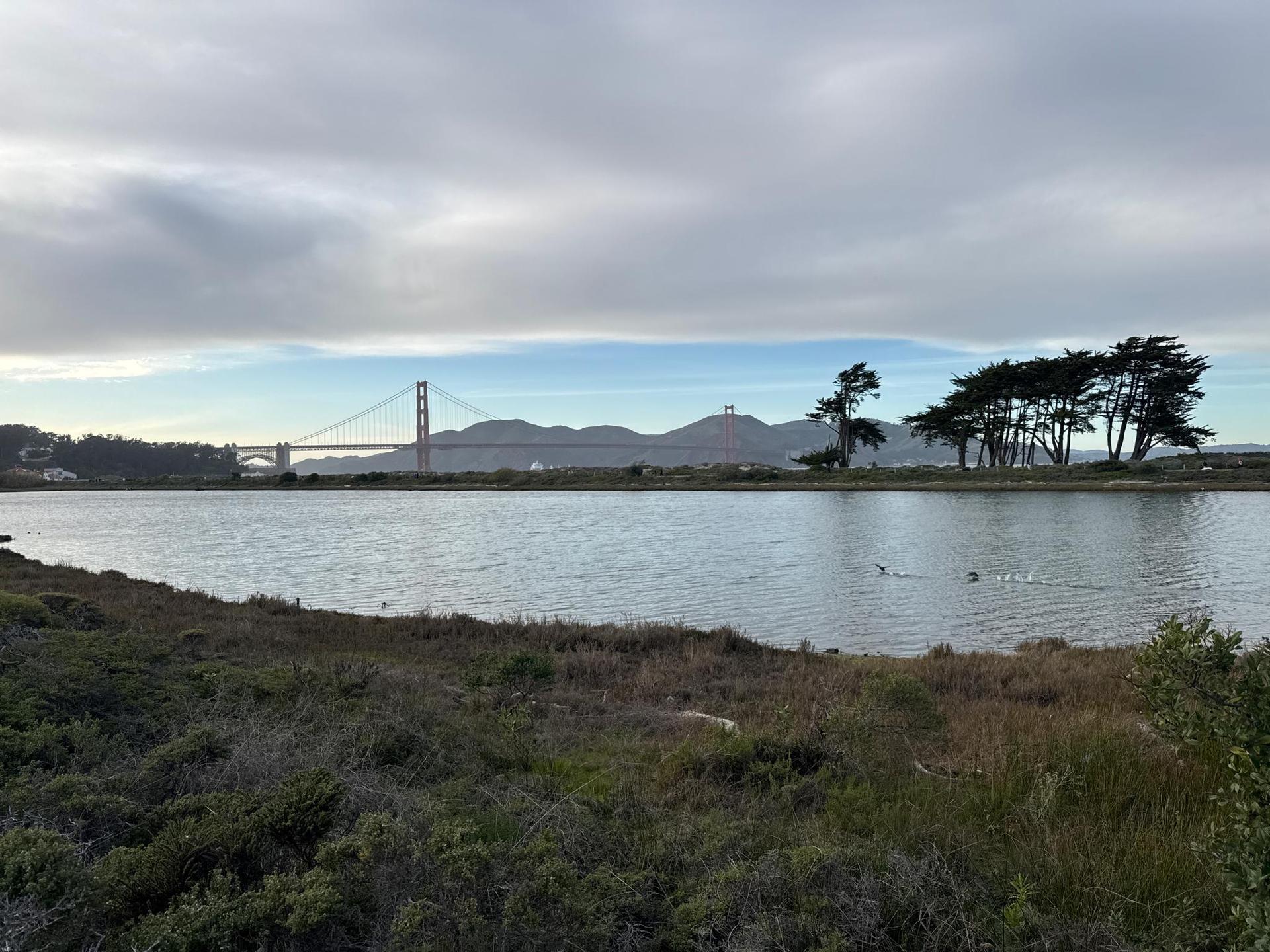 A red suspension bridge over water with distant hills under a cloudy sky.
