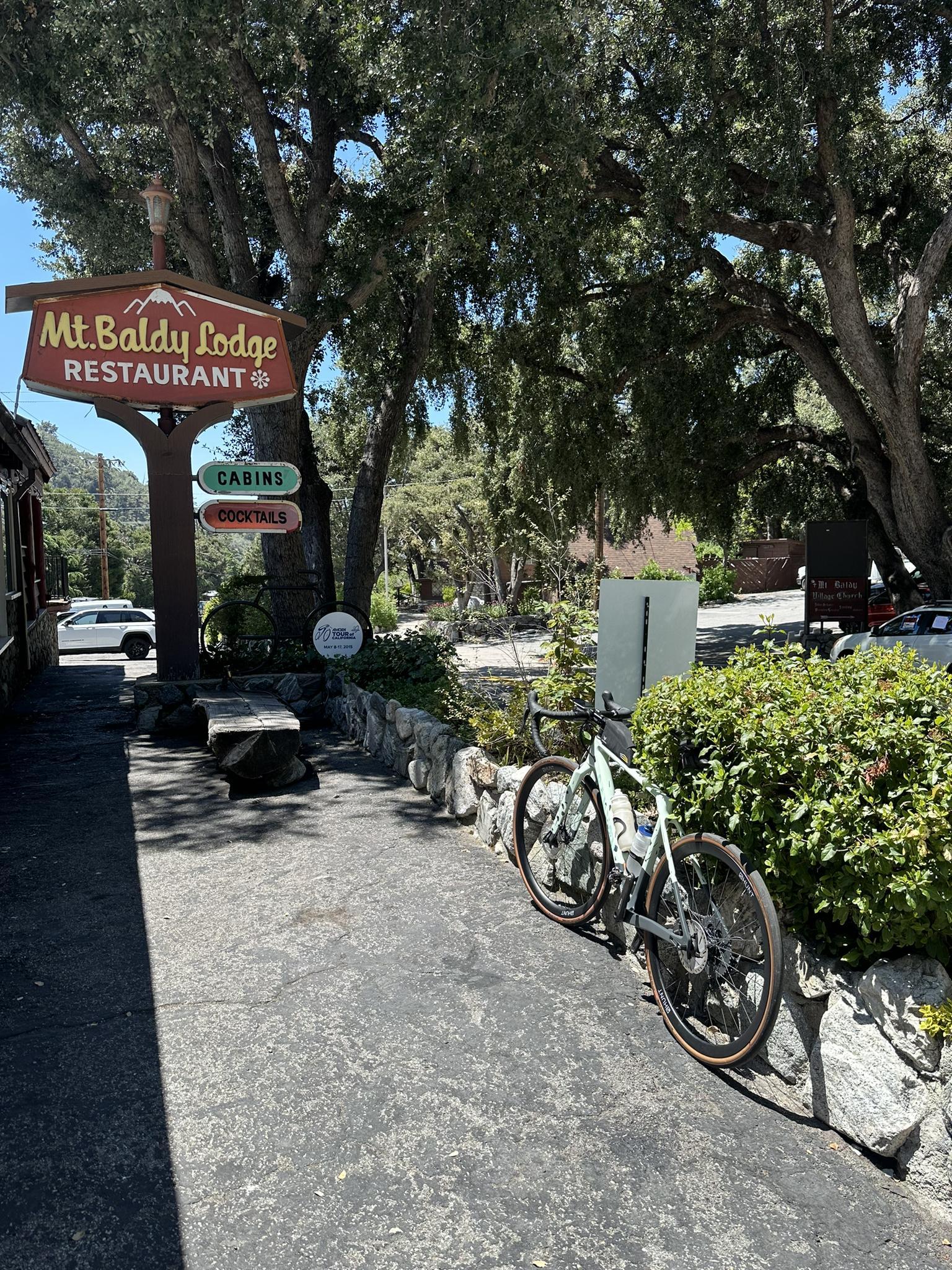 A restaurant sign under large trees, with a bicycle parked on a sunny paved path.