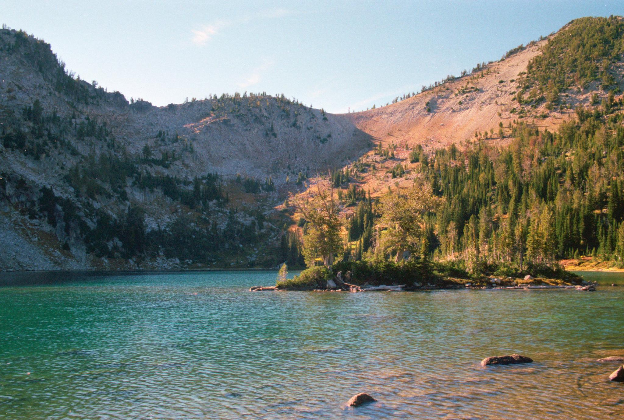 A clear mountain lake with a small island and sunlit forested peaks.