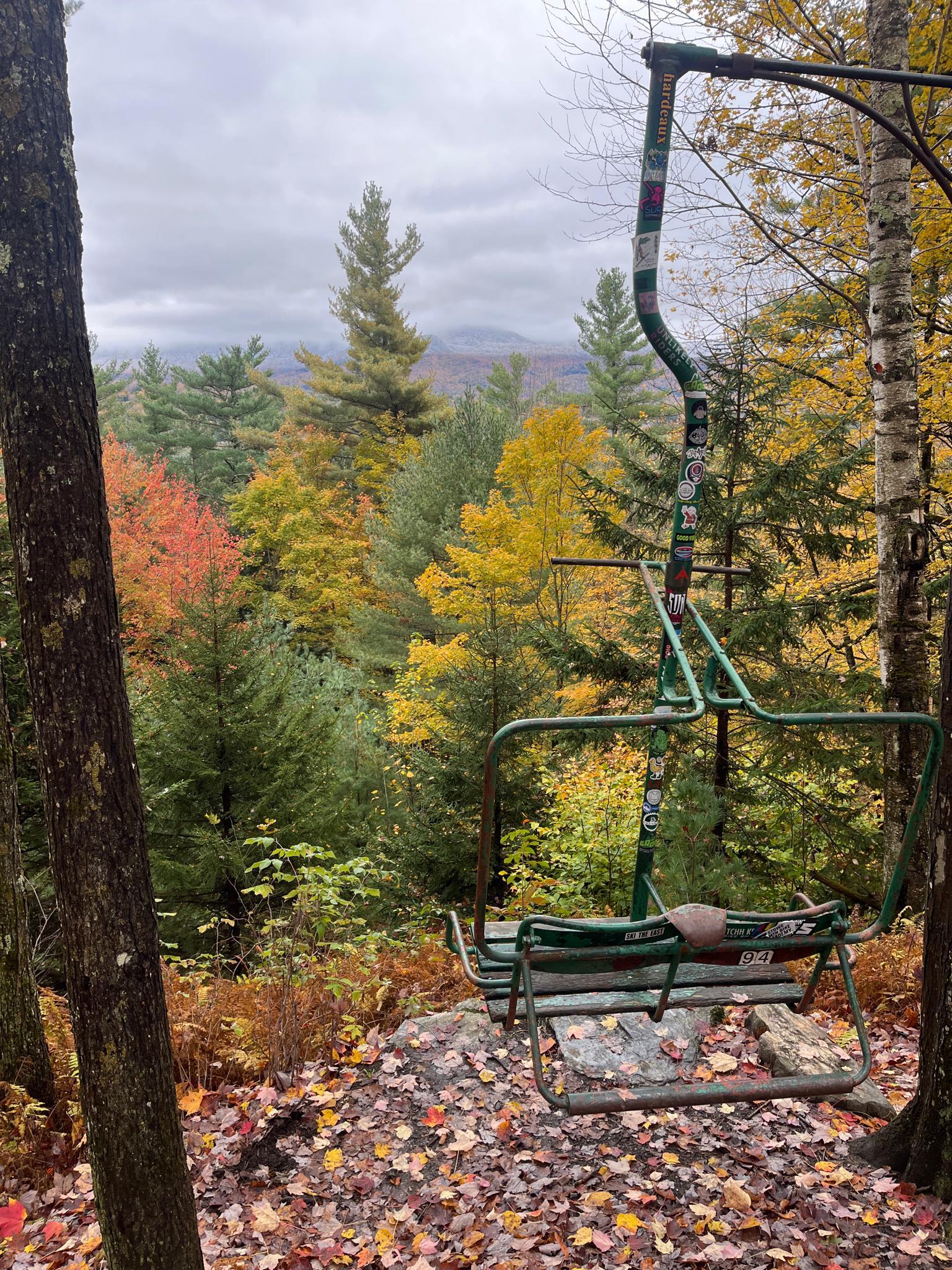 An old green ski lift chair overlooks a vibrant autumn forest and mountains.