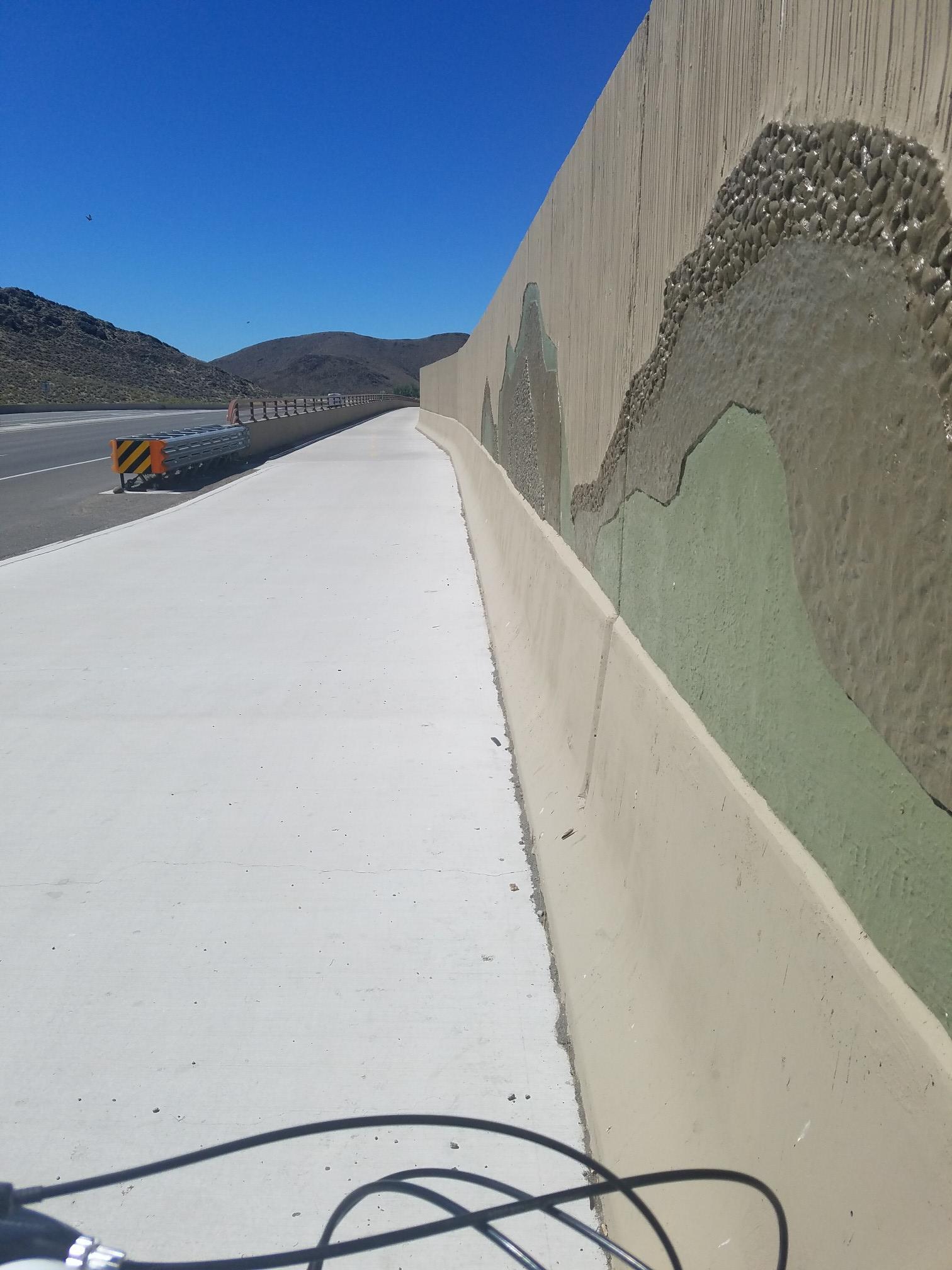 Concrete path next to a wall with mountain art, road, and distant mountains.