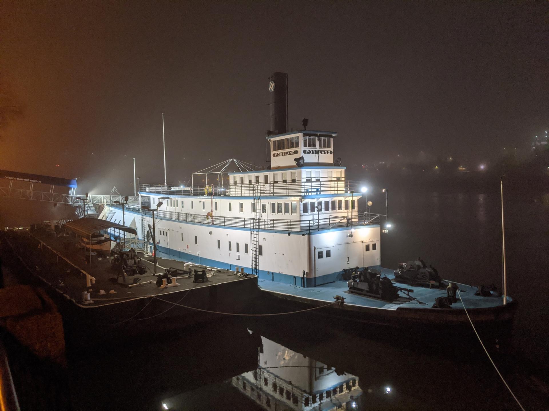 A brightly lit steamboat is docked on a foggy river at night.