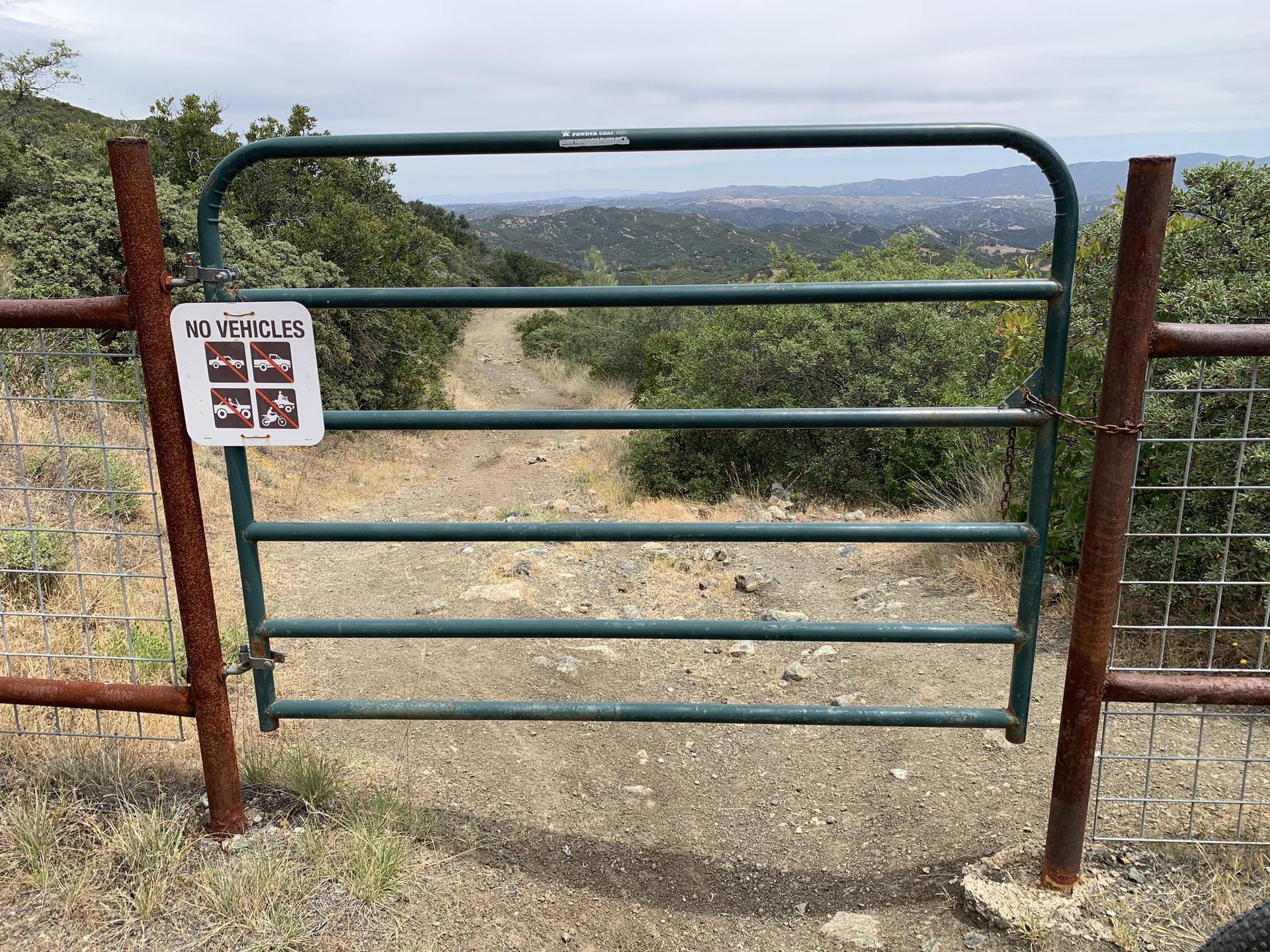 A green metal gate blocks a dirt path leading to rolling hills under a cloudy sky.