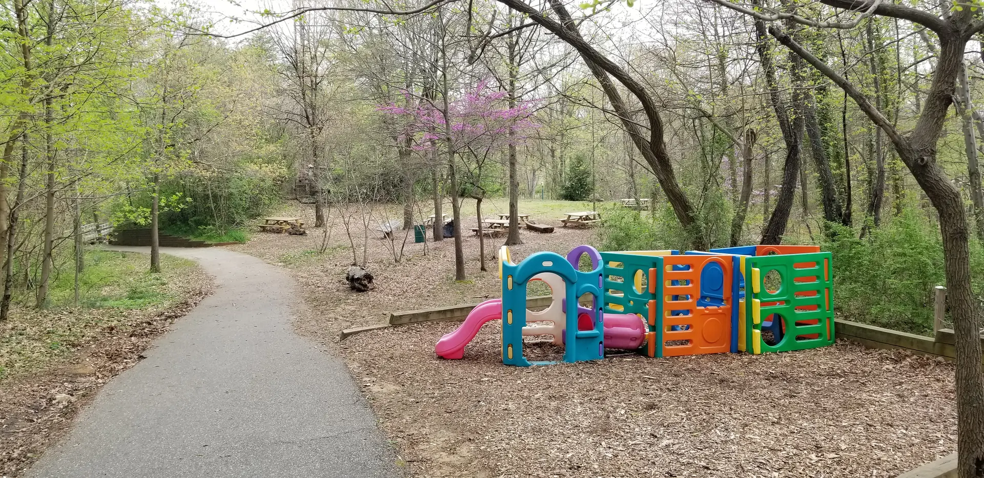 Colorful playground beside a paved path in a wooded area with picnic tables.