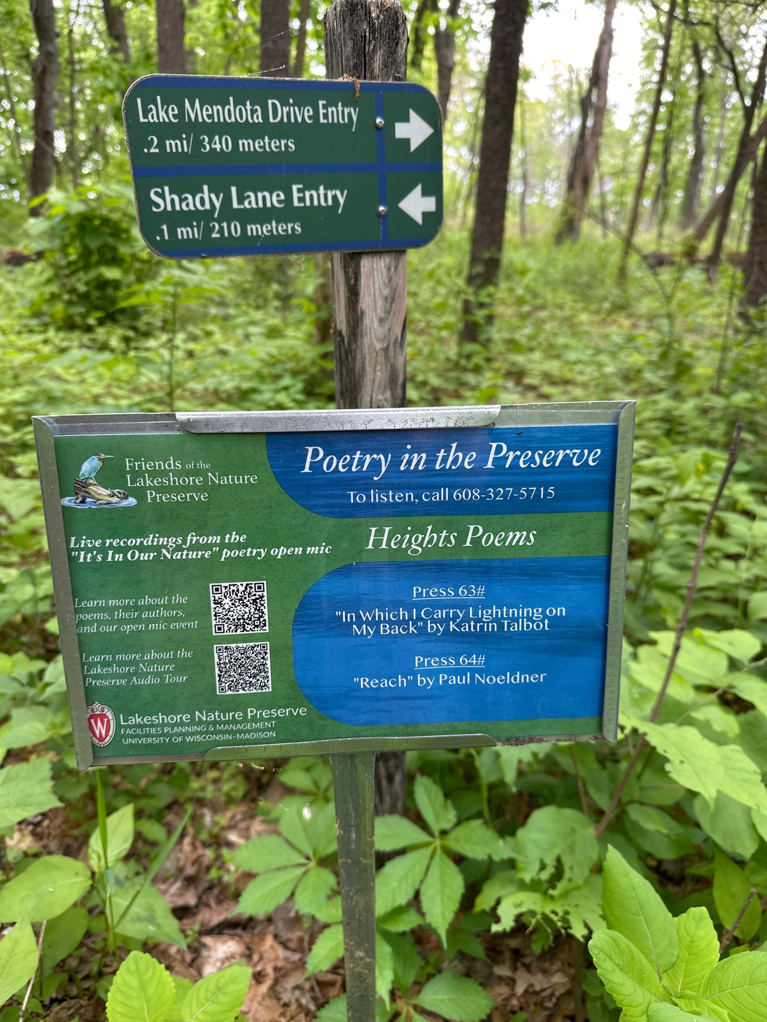 Trail signs and an informational board stand amidst lush green forest under cloudy sky.