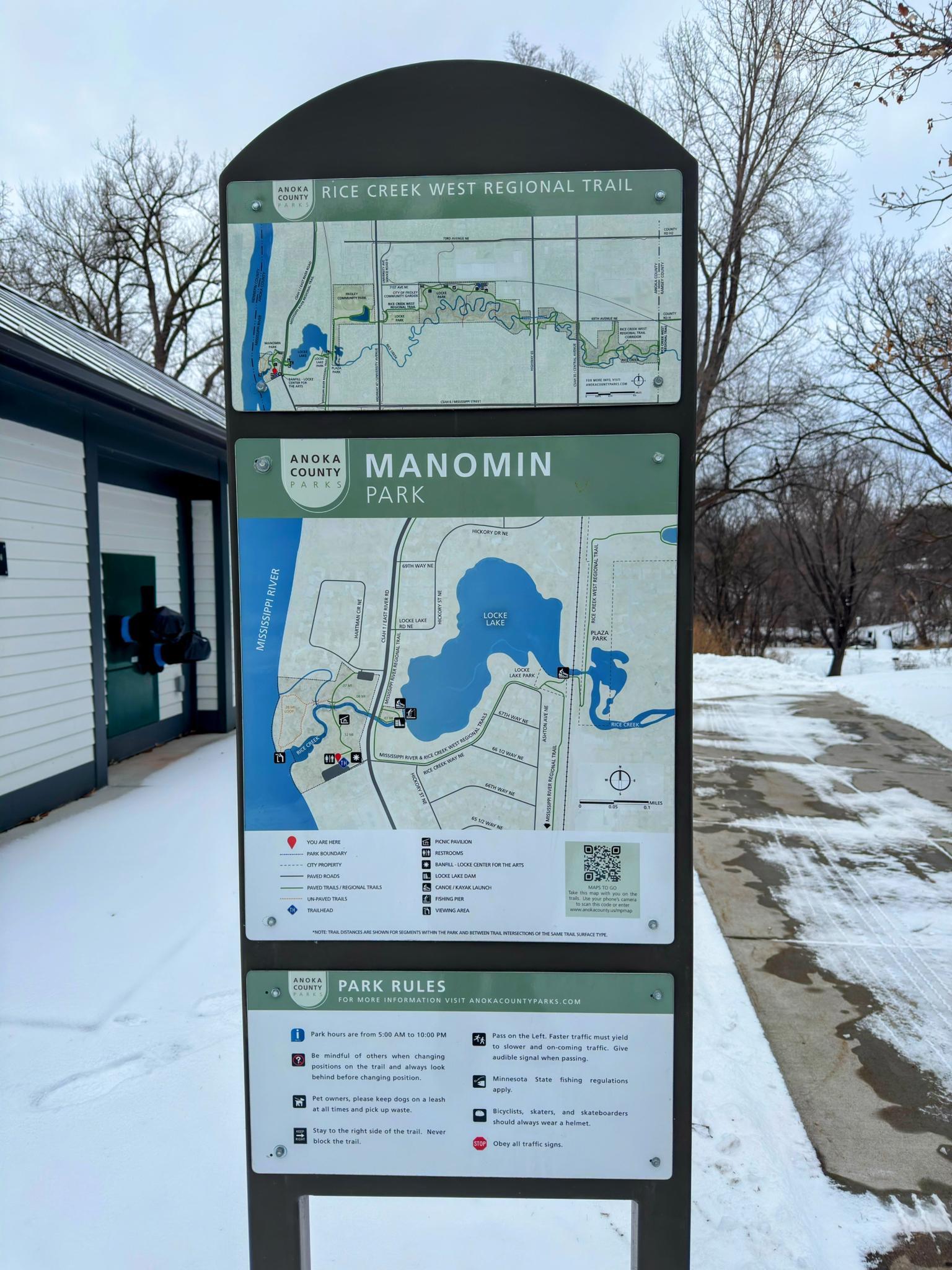 A park information sign with maps stands in a snowy landscape.