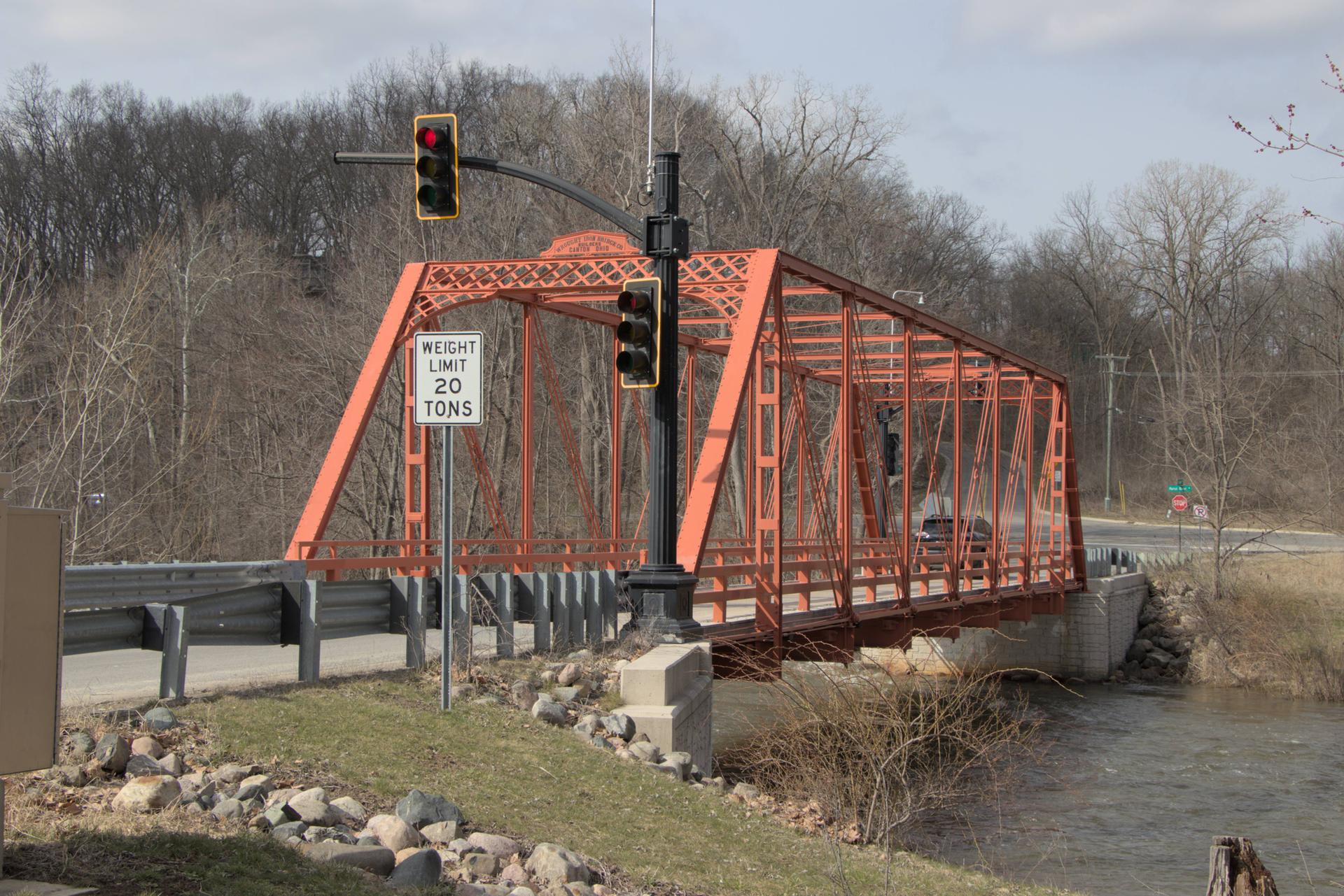 An orange metal truss bridge with a car crosses a river, bare trees in background.