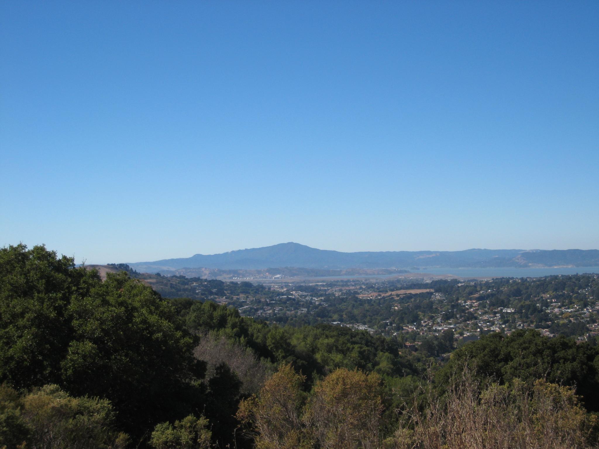 Panoramic view of a valley, urban area, distant mountains, and water under clear sky.