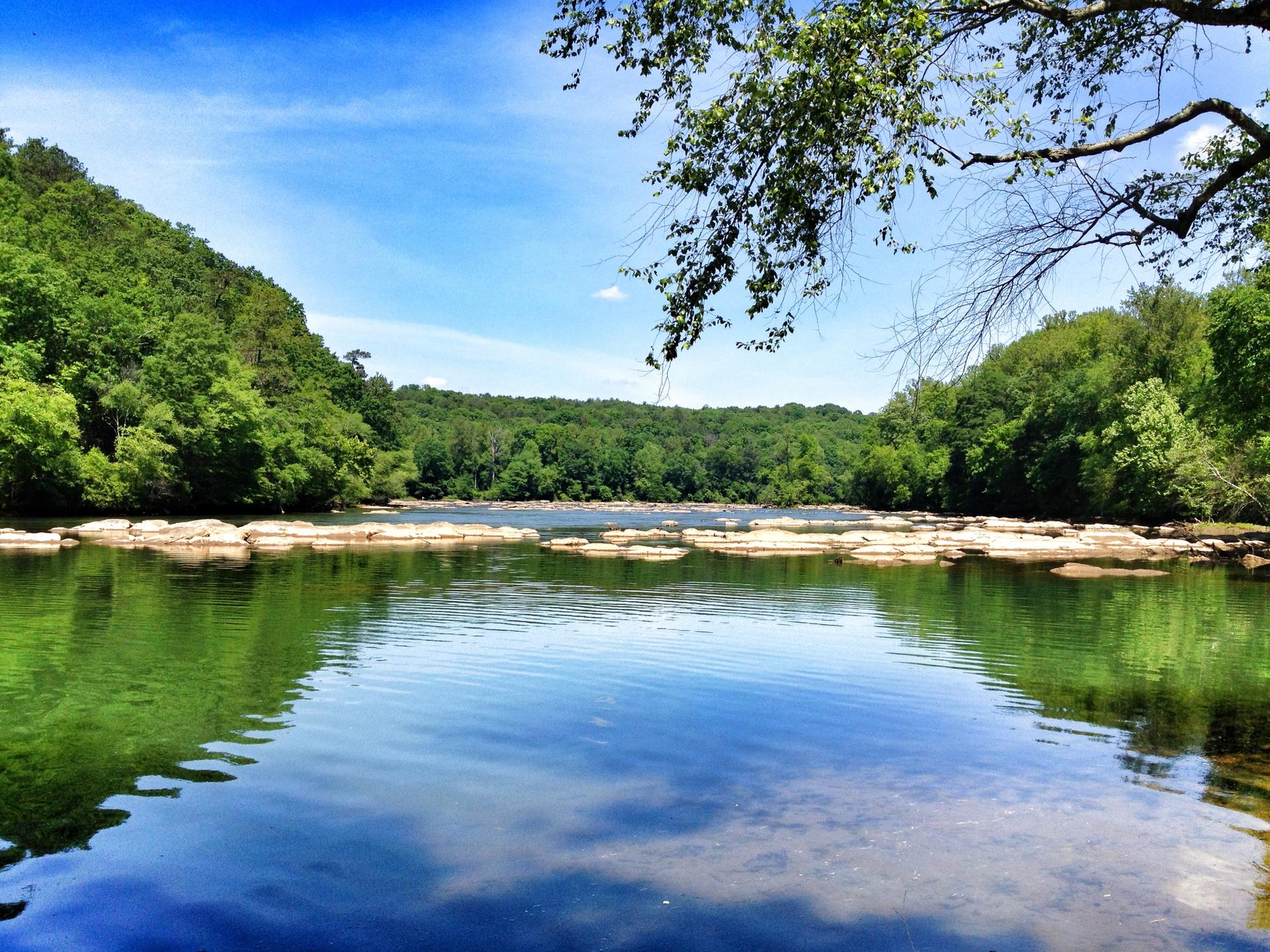 Calm river reflecting blue sky and green trees, surrounded by lush hills.