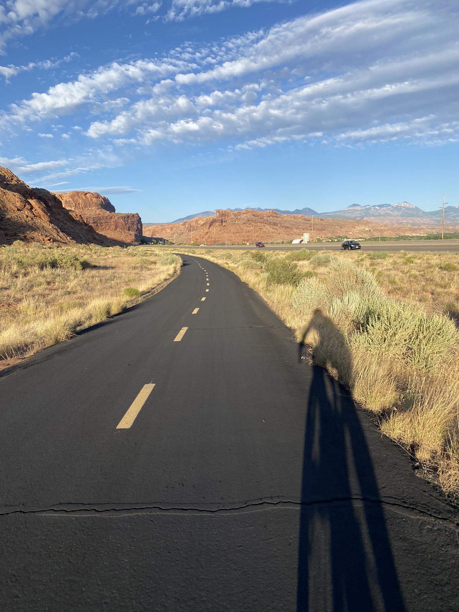 Paved road with yellow lines, long shadow, desert landscape, blue sky.