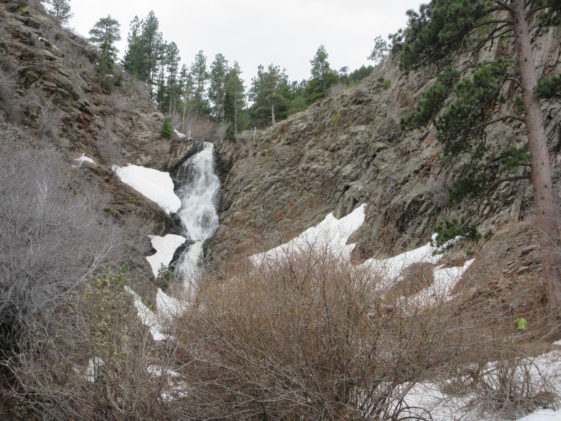 A waterfall flows down a rocky canyon with snow patches and a person.