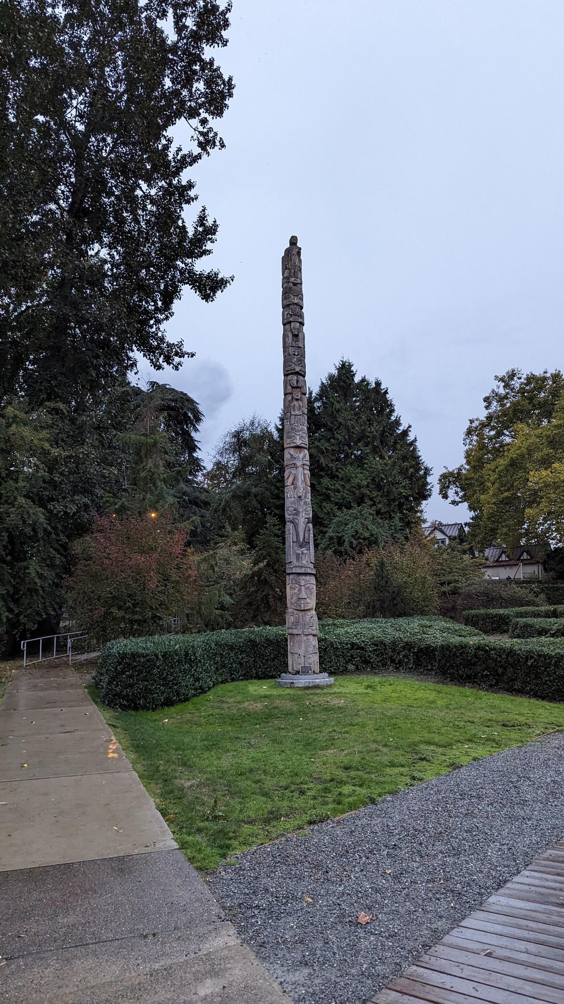 Tall, carved wooden pole standing in a grassy park area under an overcast sky.