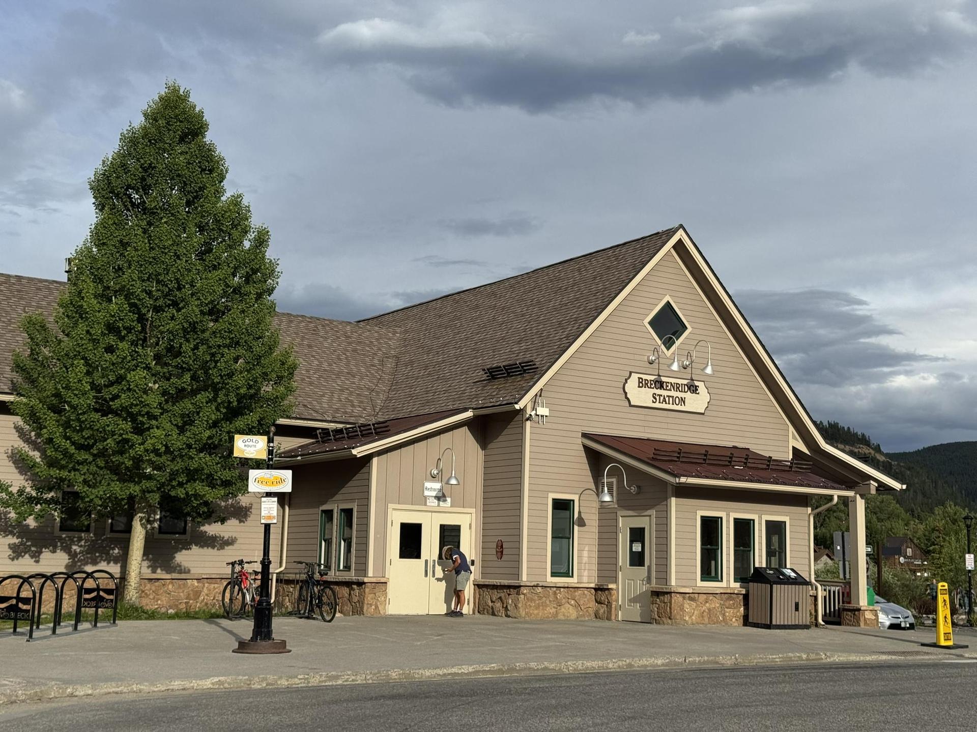 Tan building with a brown roof and large evergreen tree, person near entrance.