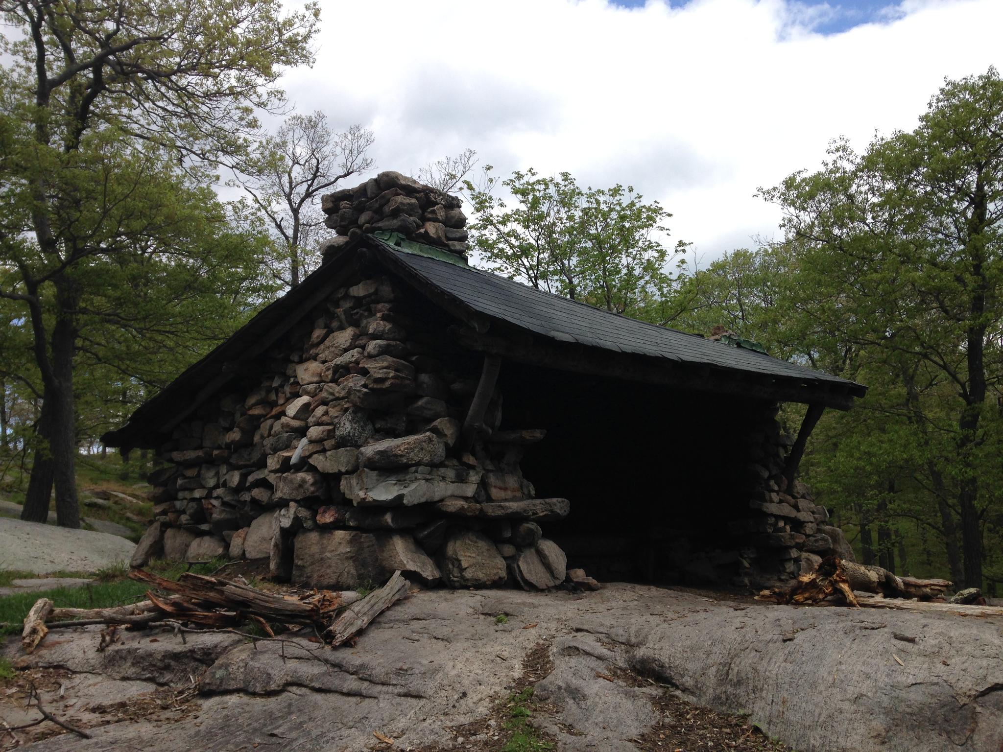 Rustic stone lean-to on a rock, surrounded by green trees under an overcast sky.