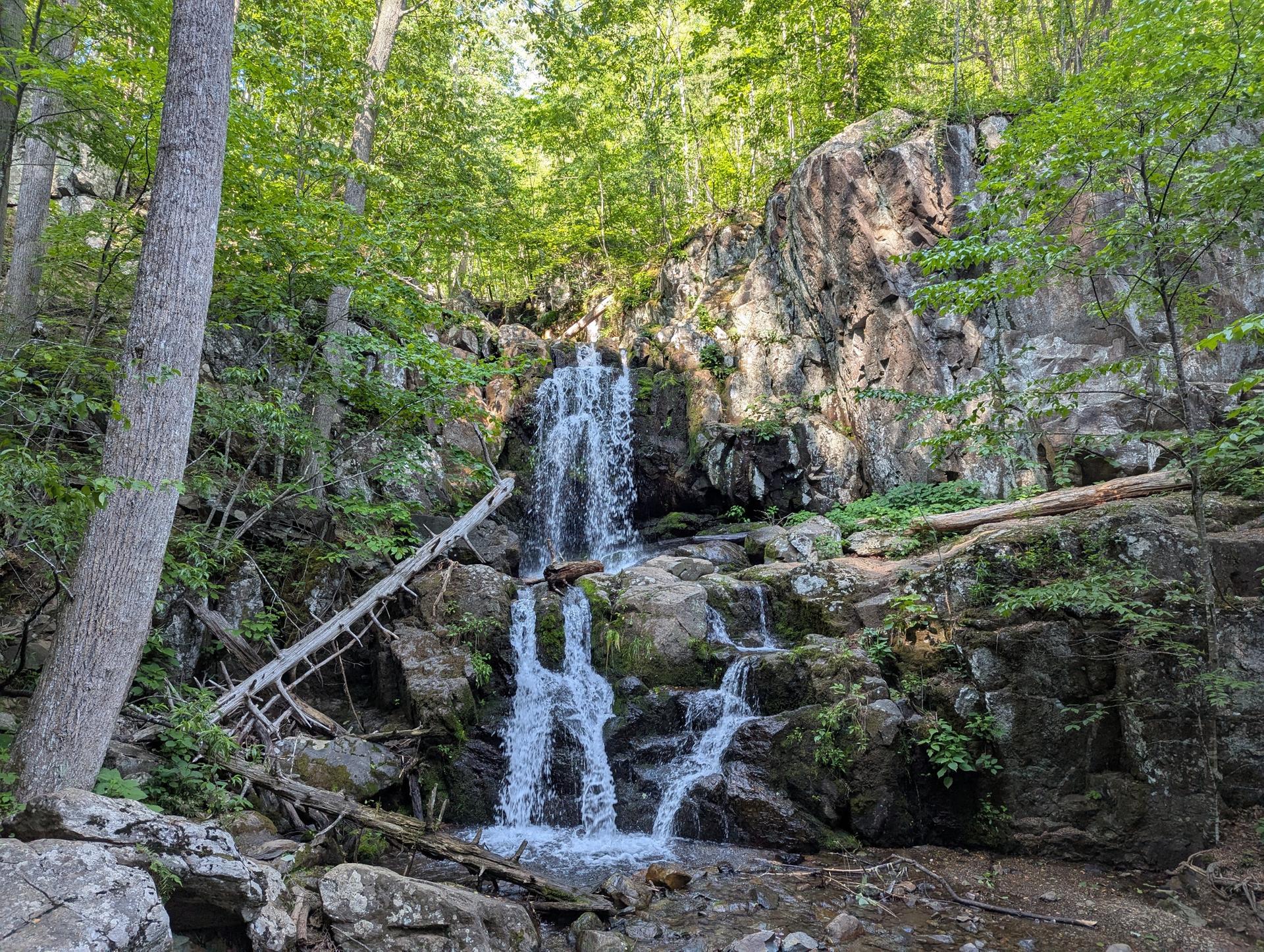 Multi-tiered waterfall flowing over large rocks in a sunlit, green forest.