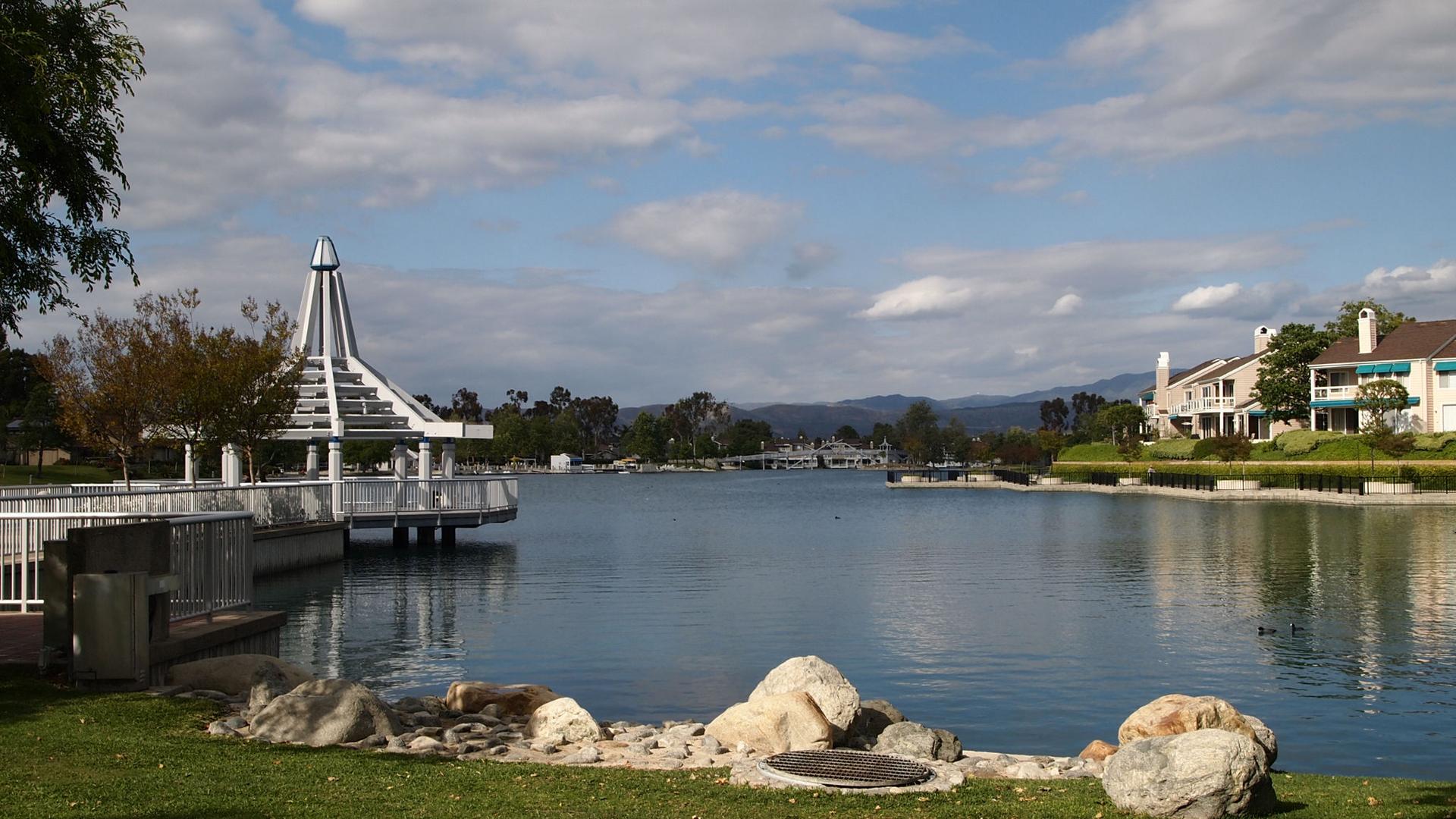 Wide view of a lake with a white gazebo, houses, and distant mountains under a cloudy sky.