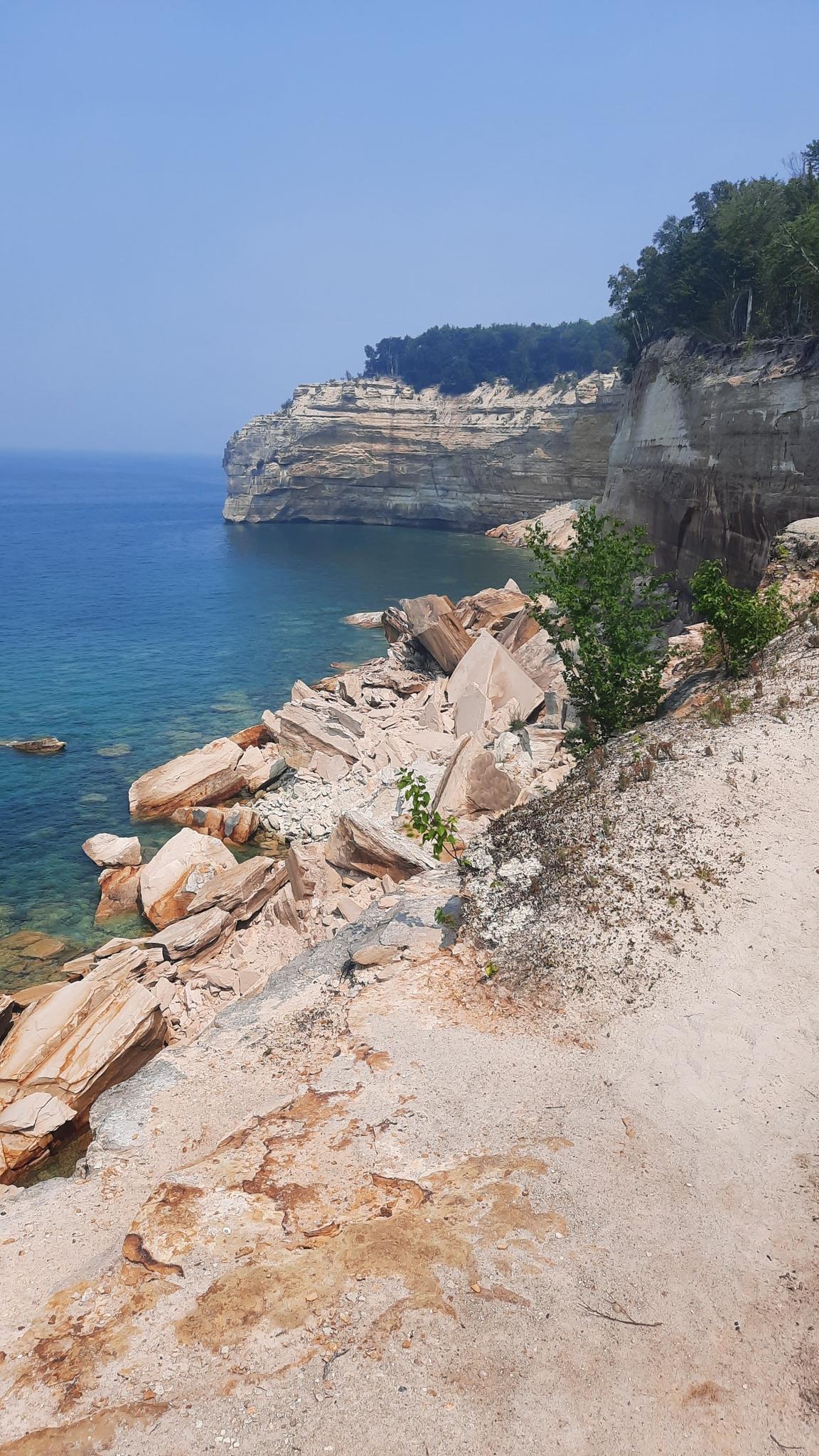 Rocky shoreline with clear blue water and a distant cliff under a bright sky.