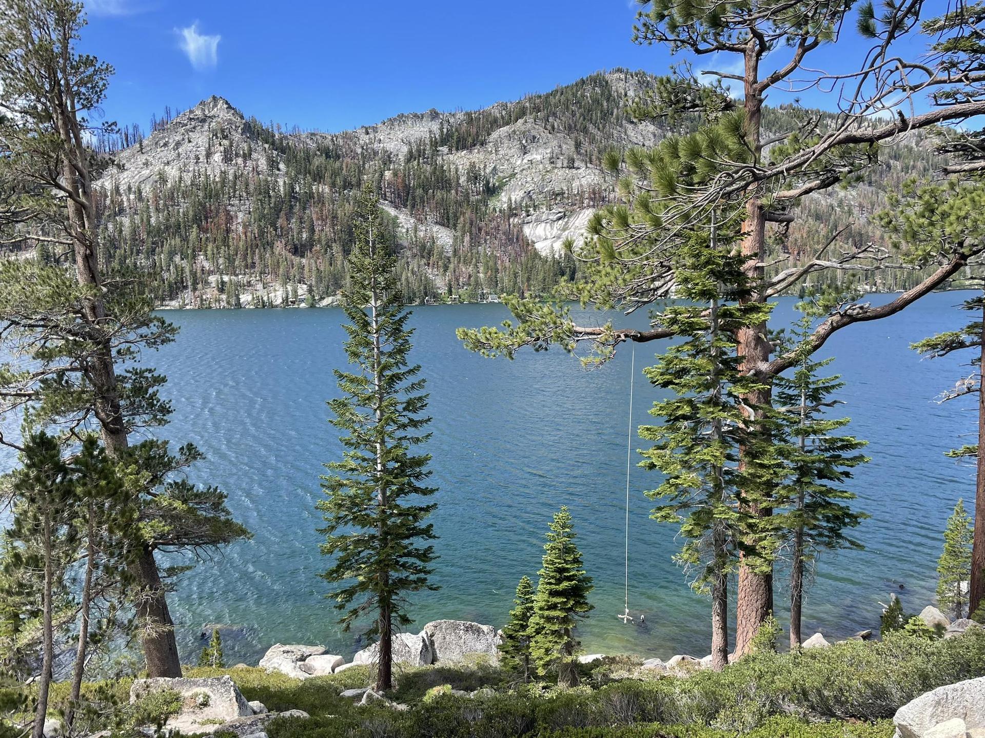 Clear blue lake surrounded by rocky mountains and pine trees, rope swing visible.