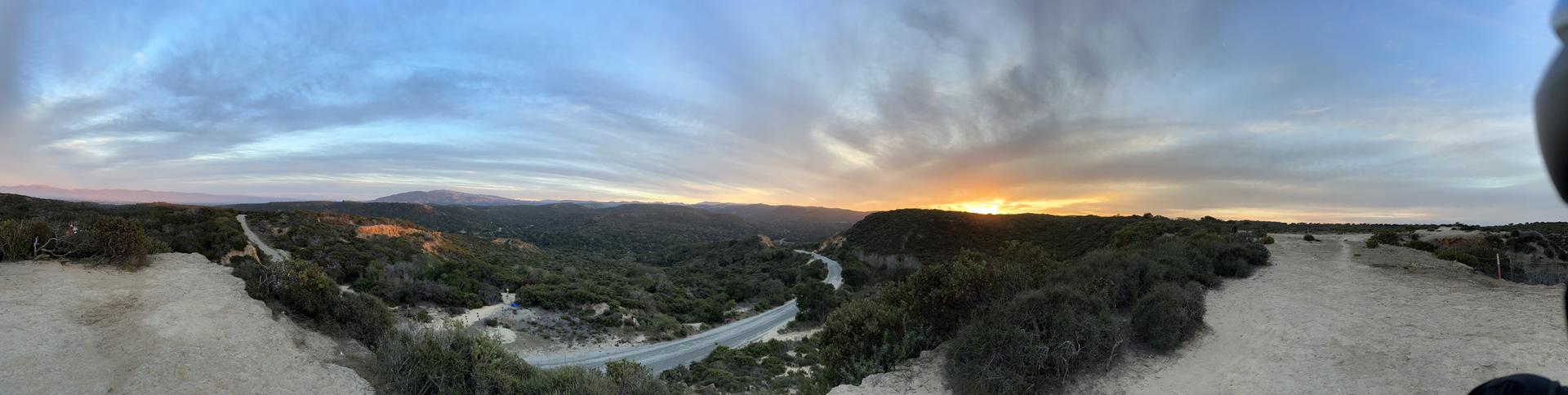 Panoramic sunset over a valley with a road and dense trees.