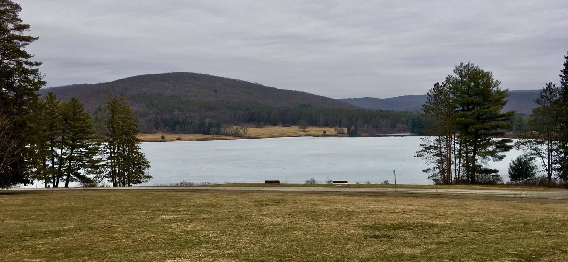 Overcast view of a calm lake bordered by grassy banks and distant forested hills.