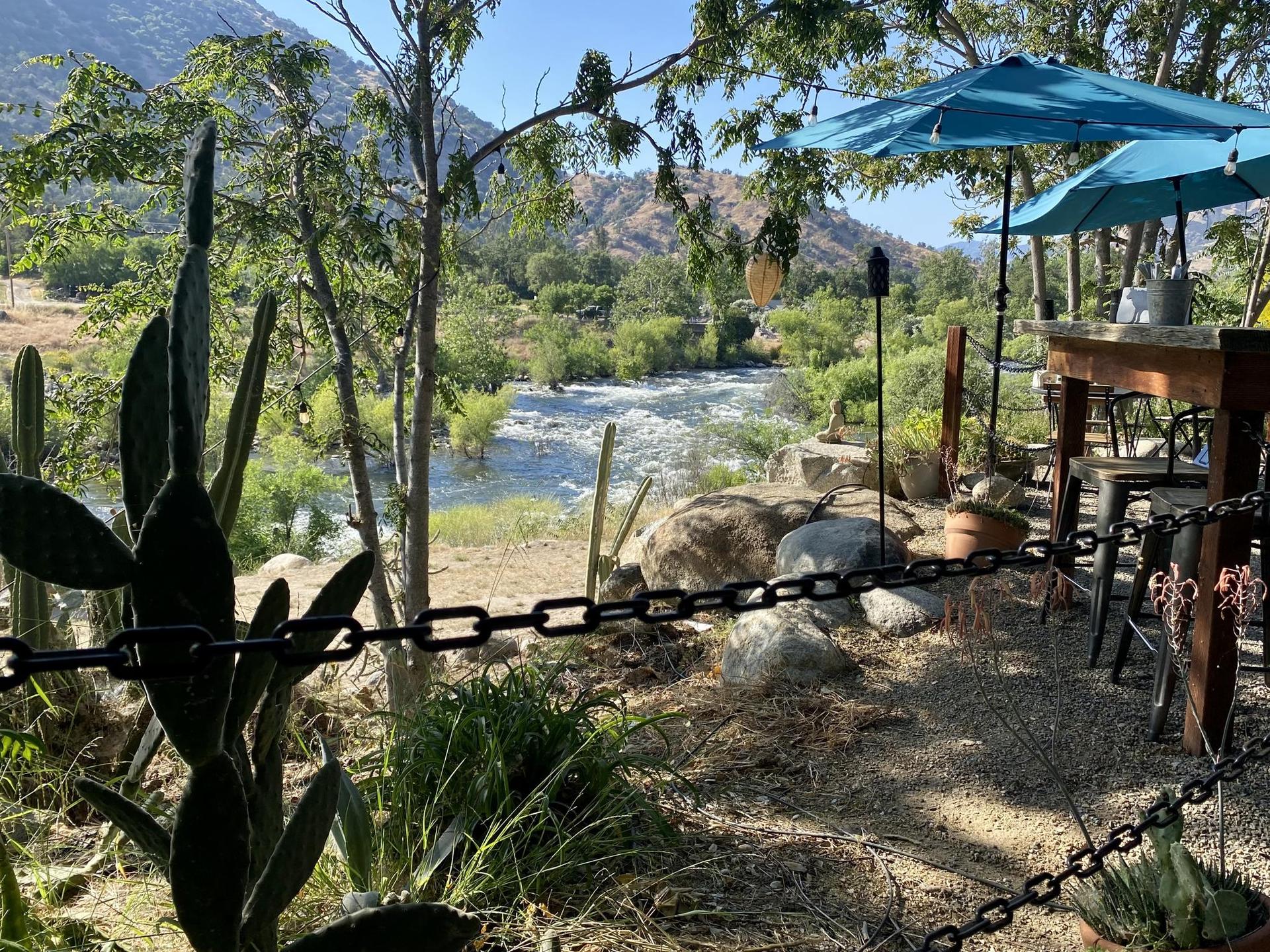 Sunny outdoor seating area with cacti overlooks a flowing river and green mountains.