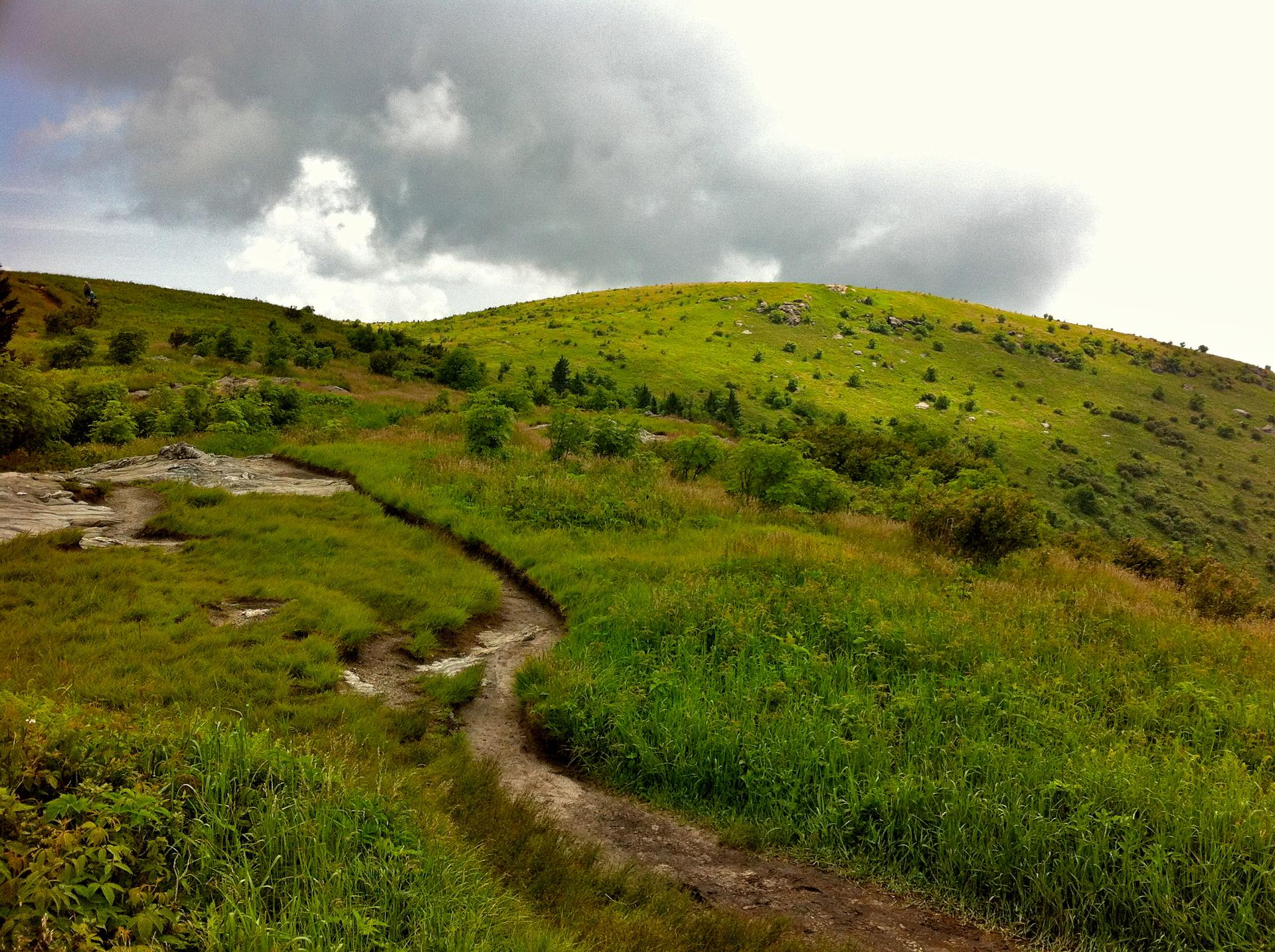 Winding dirt path through vibrant green hills under a dramatic cloudy sky.