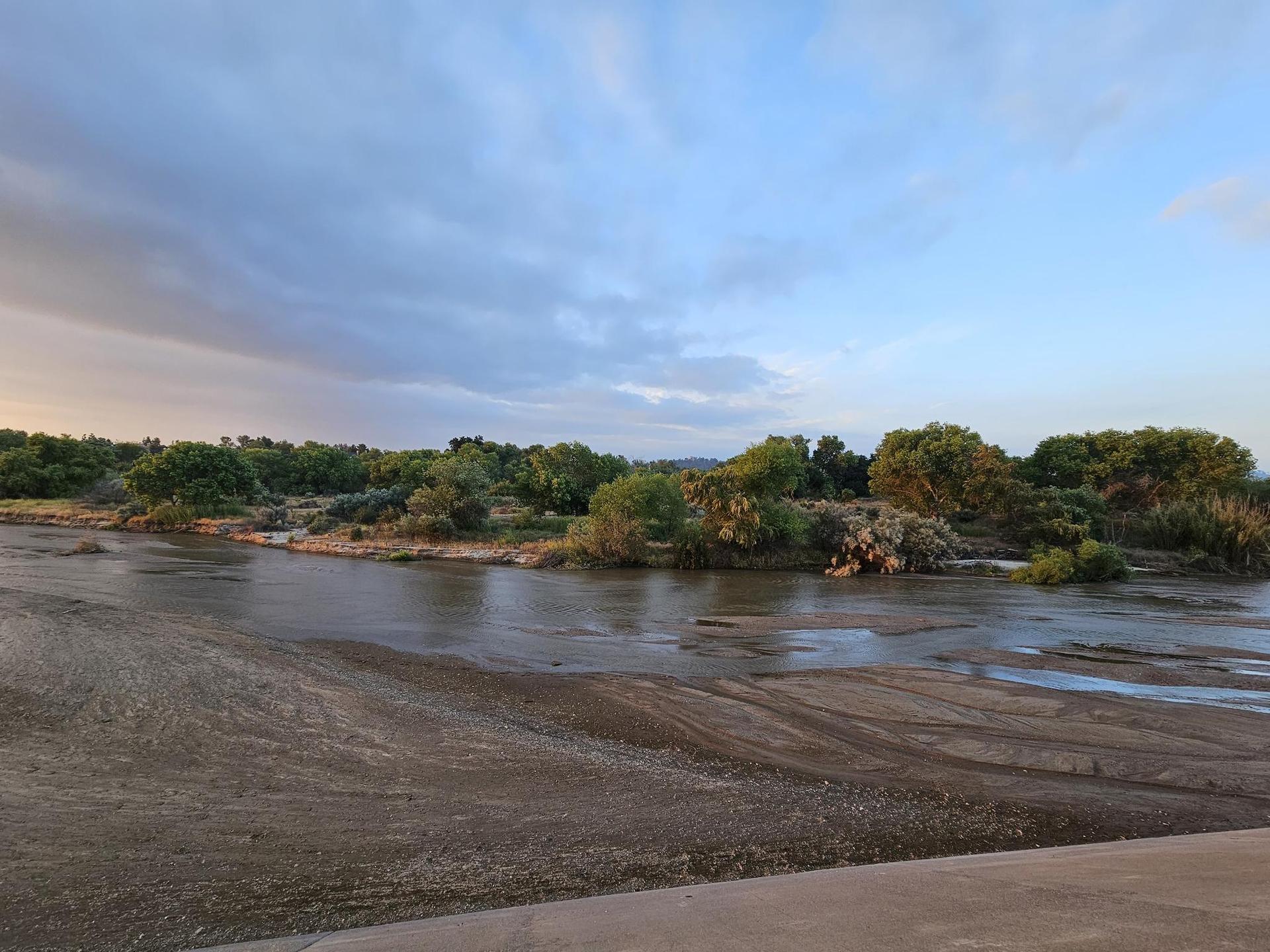 A wide river with muddy banks and lush green trees under a cloudy sky.