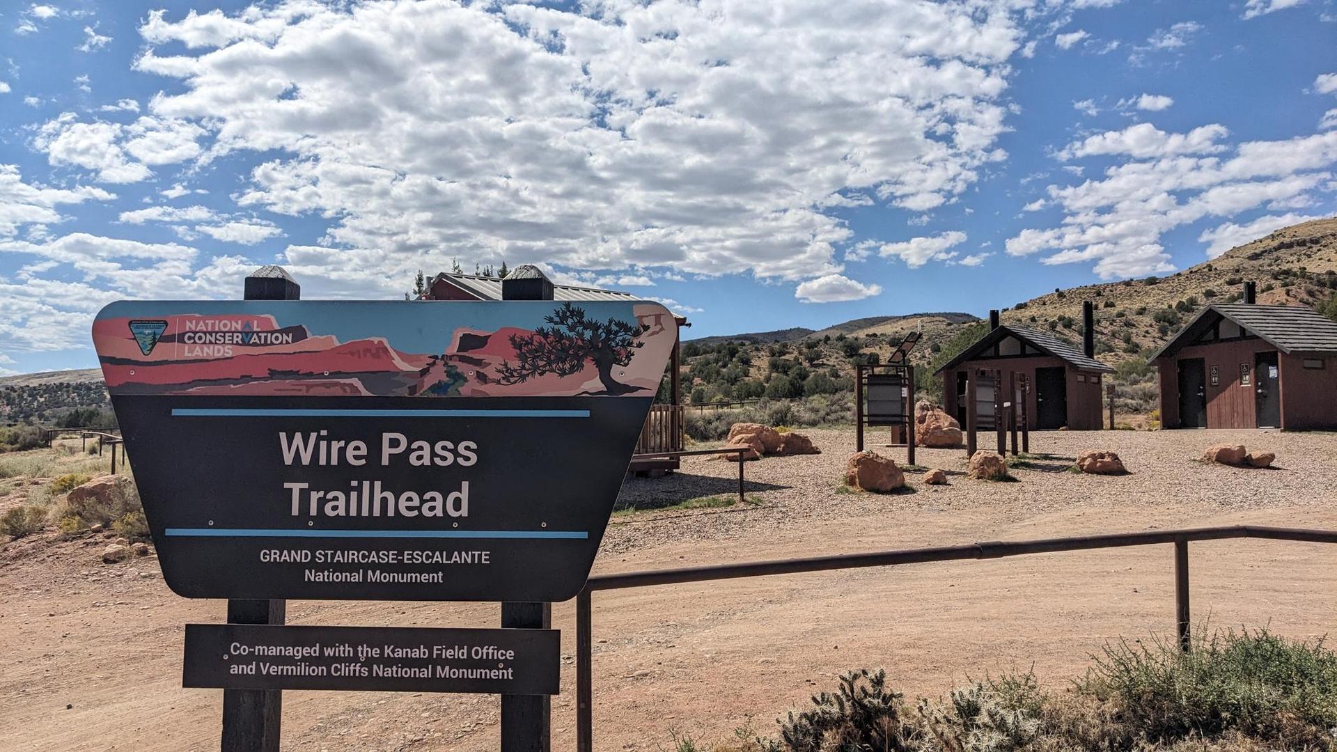 Trailhead sign for "Wire Pass" with small wooden buildings under cloudy sky.