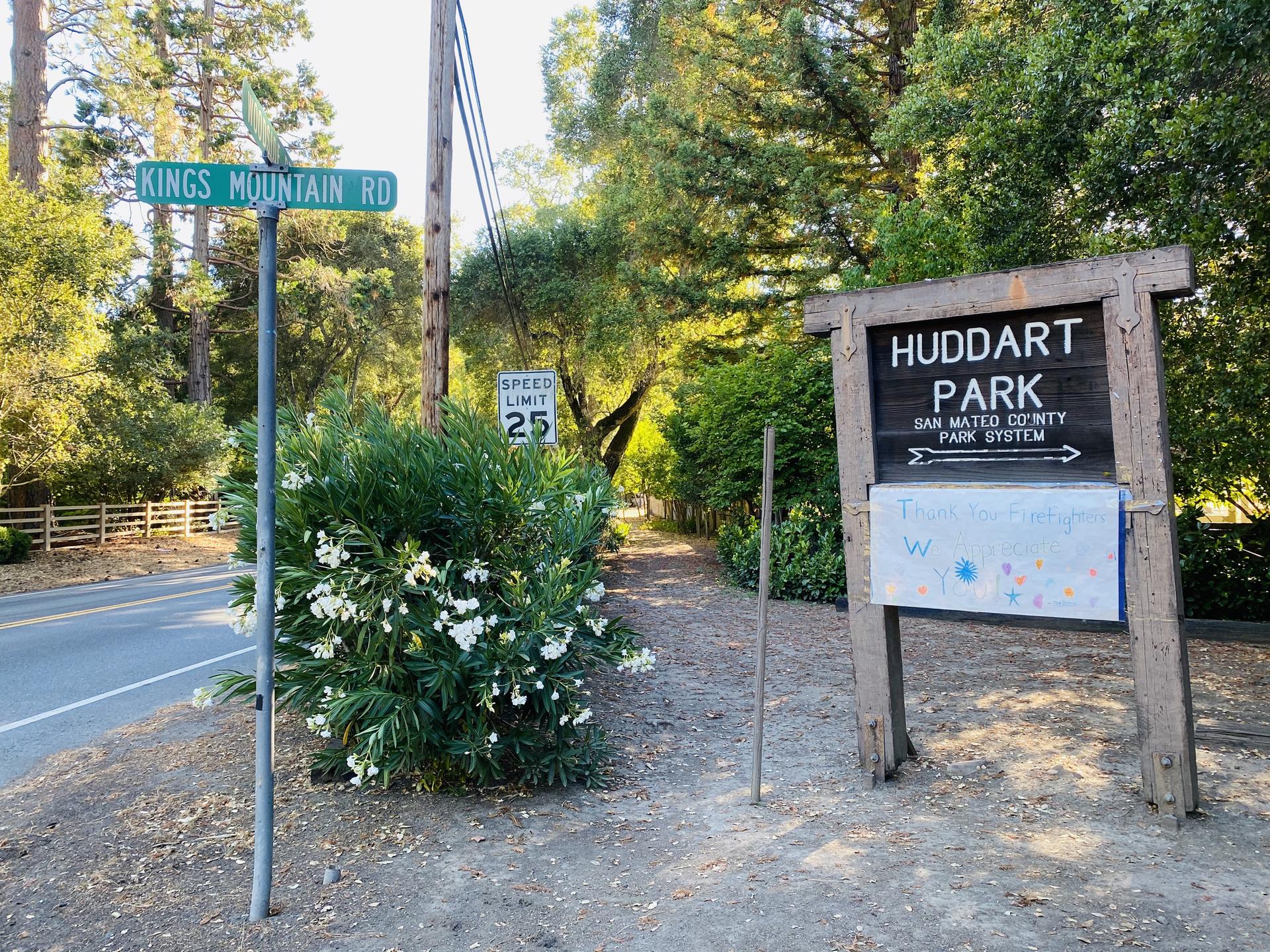 Road sign and wooden park sign next to a road and green bushes.