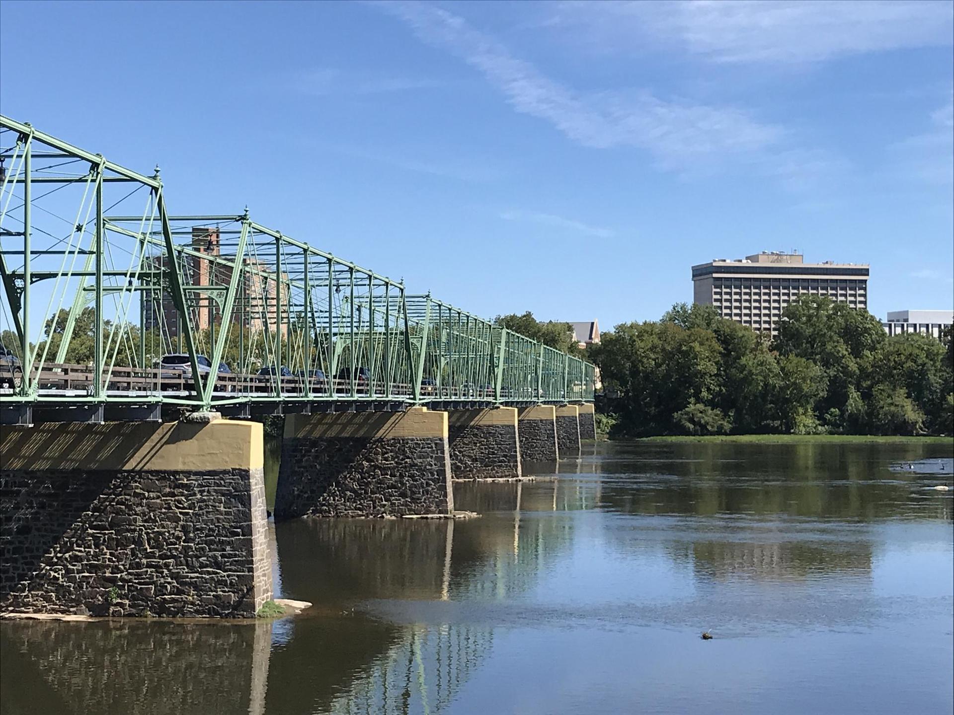 A long green metal truss bridge spans a wide river on a sunny day.