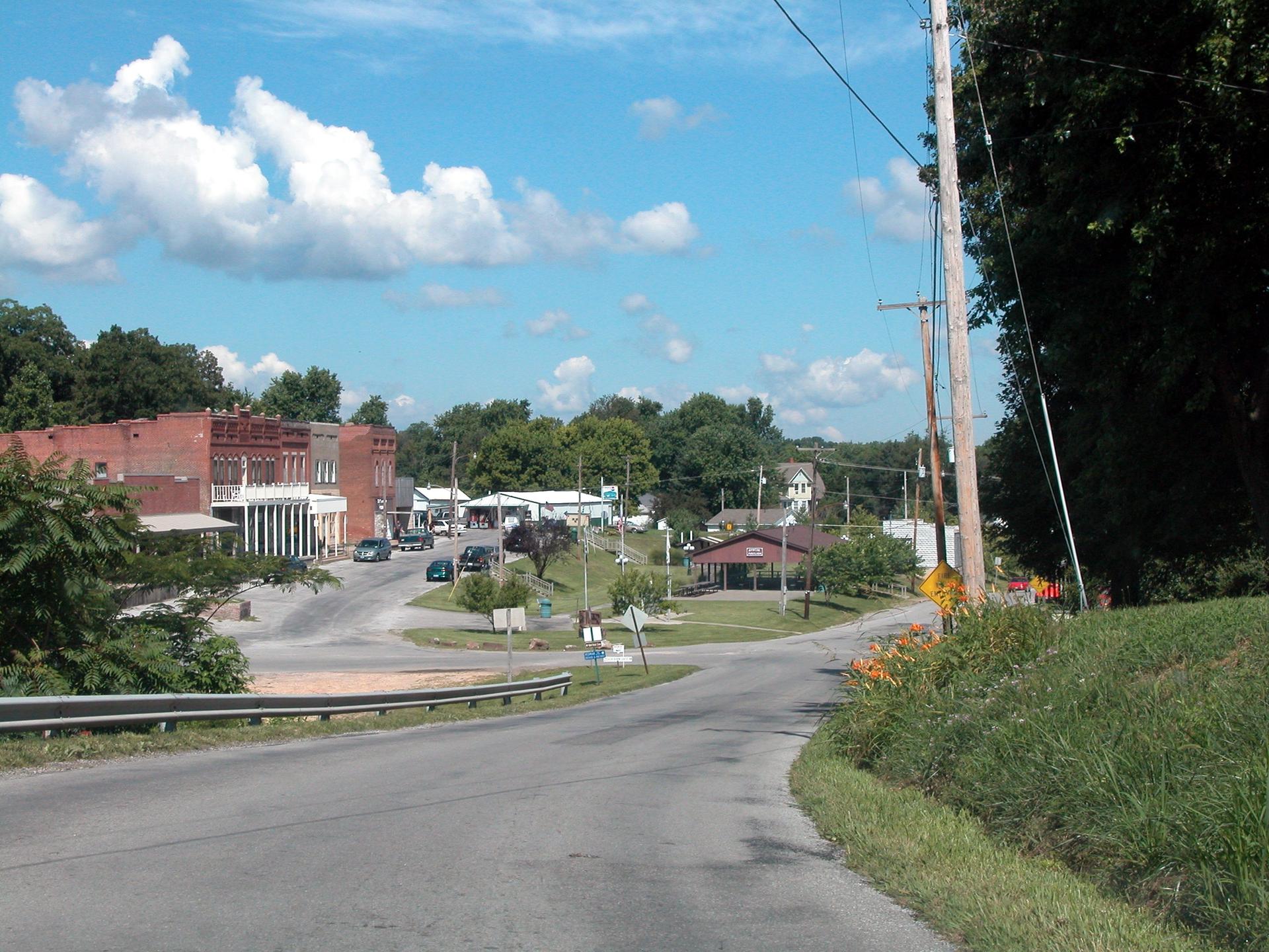 Sunny view of a small town intersection with brick buildings and green trees.
