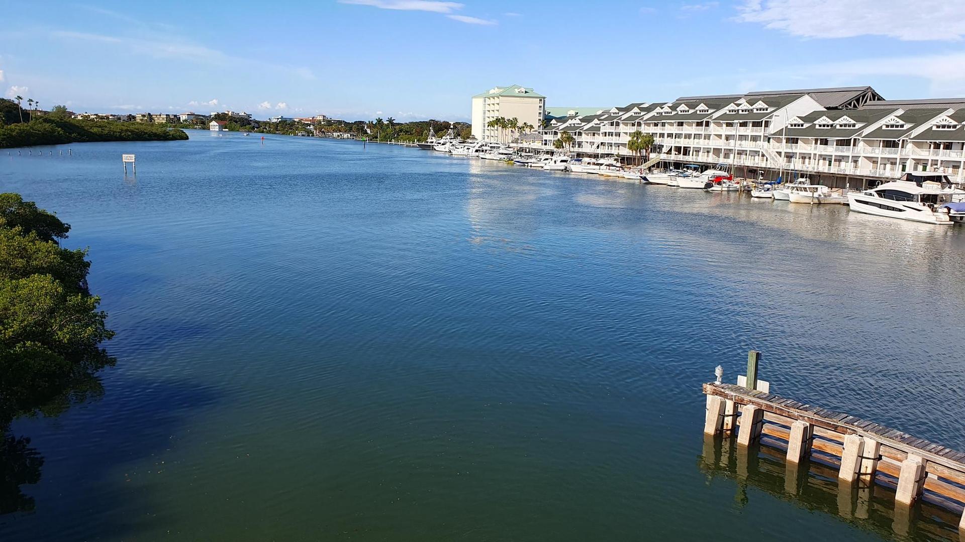 Boats docked at a marina with buildings along a wide waterway, sunny.