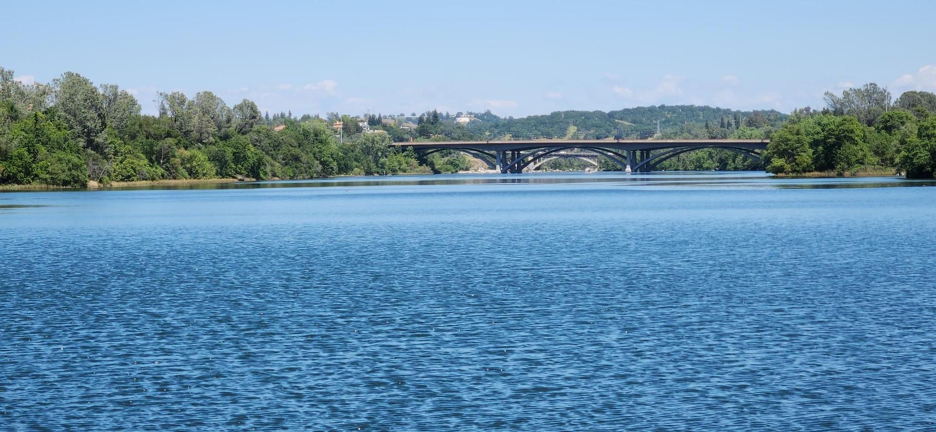 Wide blue lake with a distant bridge and green tree-lined shores.