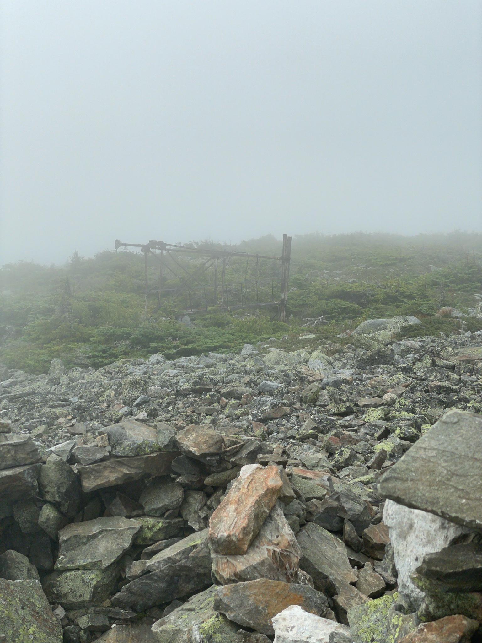 A rocky, foggy mountain landscape with a distant wooden structure and sparse greenery.