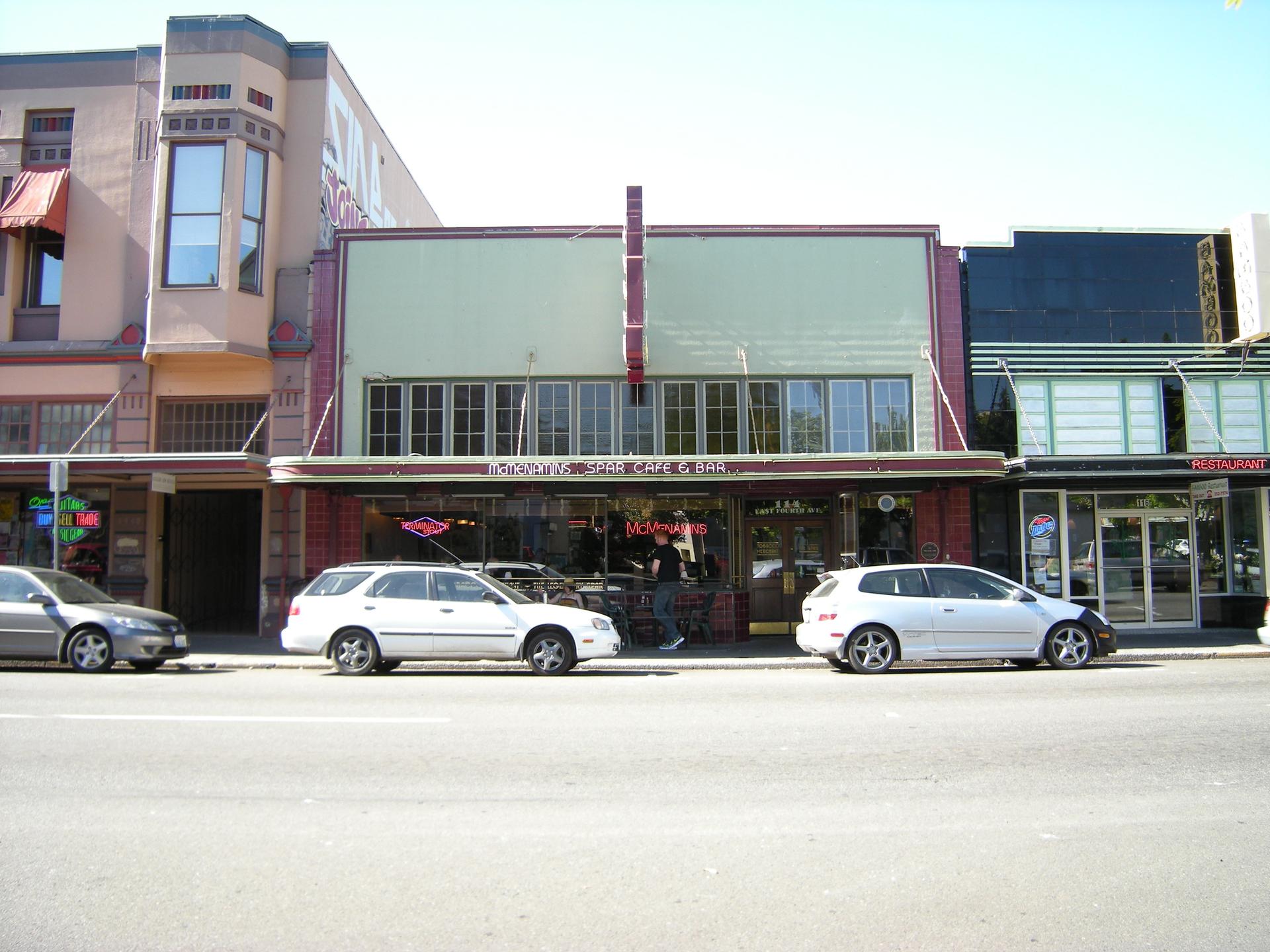 A row of urban storefronts with parked cars under bright daylight.
